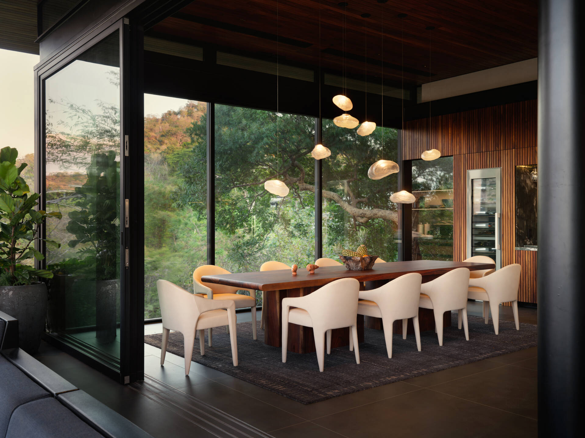 Floor-to-ceiling glass, sculptural lighting, and a long timber table create a dining space immersed in the tropical landscape.