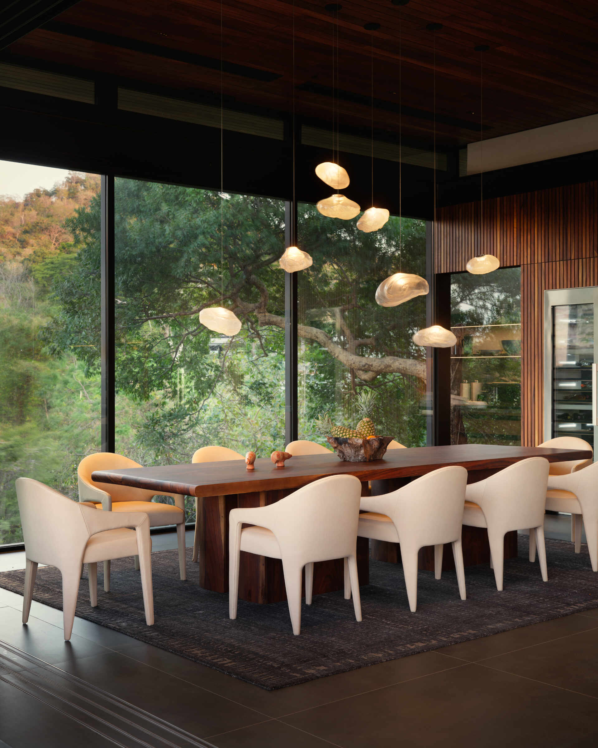 Floor-to-ceiling glass, sculptural lighting, and a long timber table create a dining space immersed in the tropical landscape.