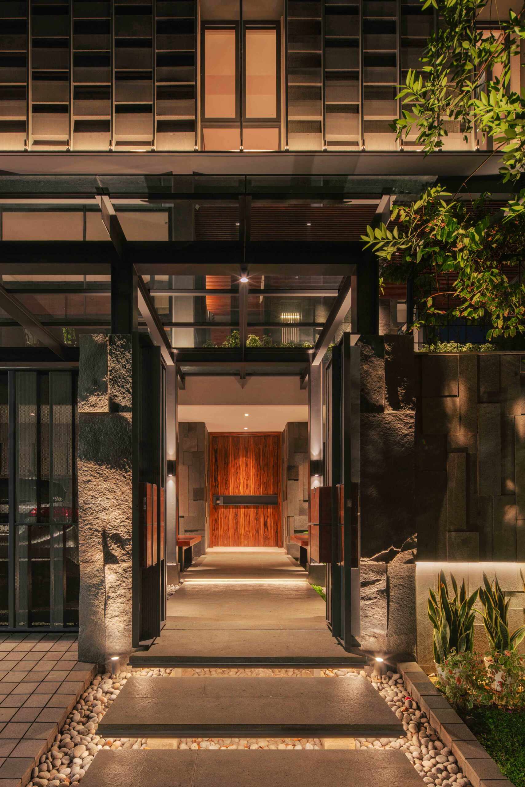 Step inside through a steel-framed entrance that leads into a landscaped stone entryway with views toward the central courtyard.