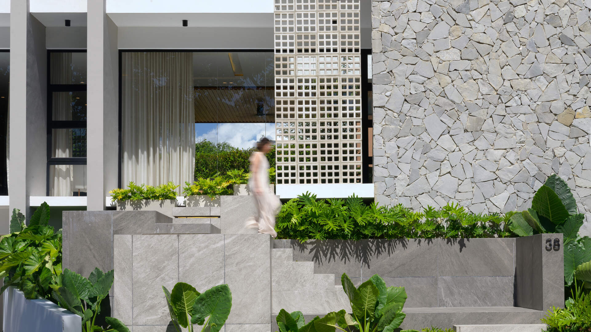 See how broad stone stairs, tropical landscaping and textured materials create a welcoming entrance to this modern hillside home.