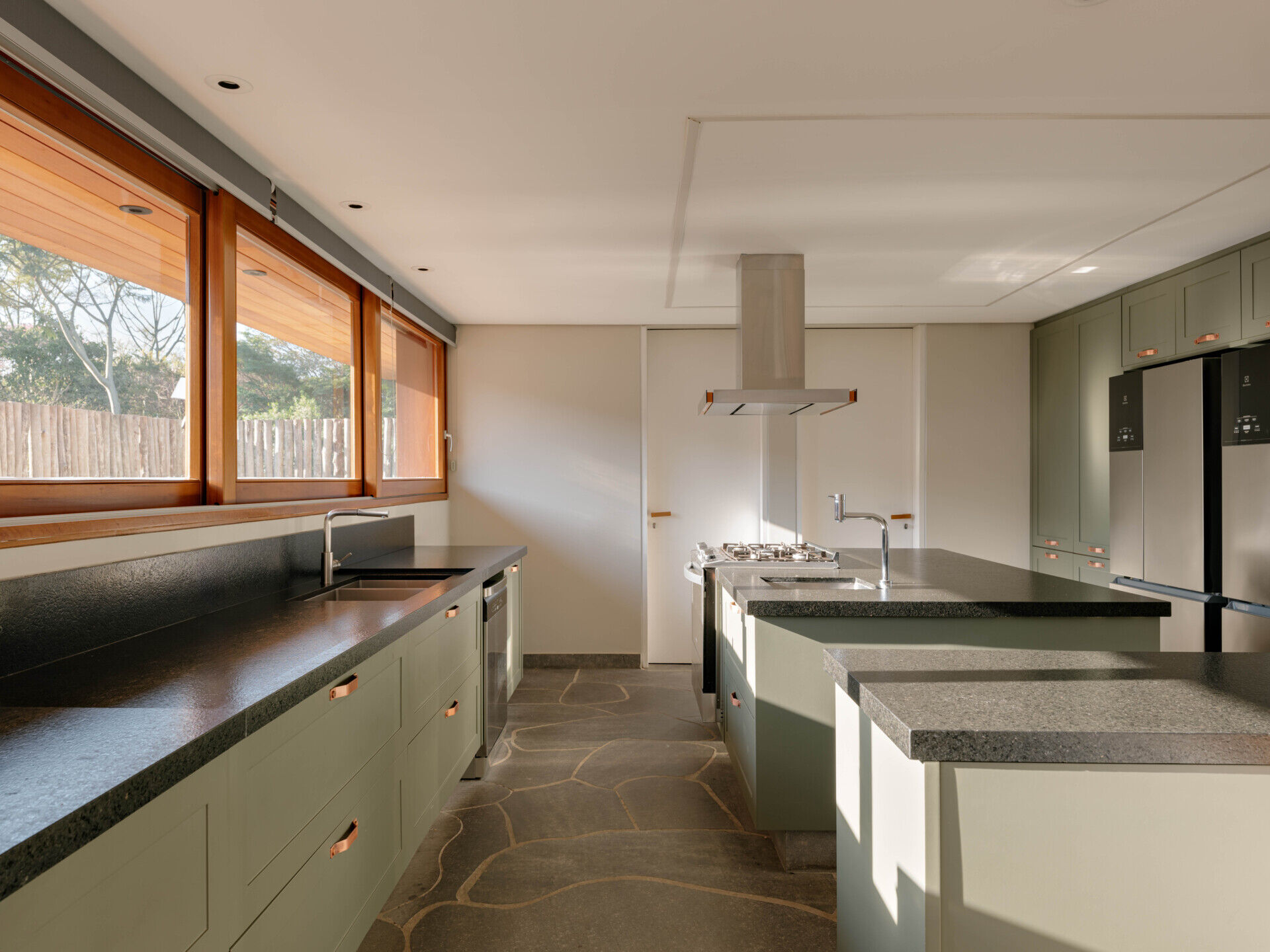 Soft green cabinetry, dark stone benchtops, basalt flooring and timber-framed windows give this contemporary country kitchen a warm, welcoming feel.