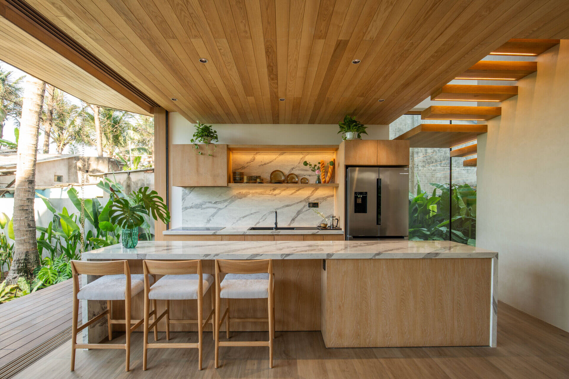 This warm, open kitchen pairs timber cabinetry, marble surfaces and a generous island with garden views, creating a bright space that flows naturally into the surrounding tropical landscape.