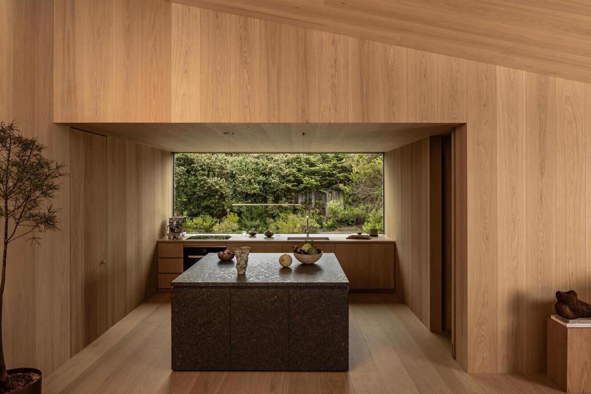 The kitchen continues the restrained palette while introducing contrast through a dark stone island. Against the surrounding oak, the textured stone immediately becomes the visual anchor without overwhelming the room.
A wide window above the worktop brings in natural light and provides another uninterrupted view across the landscape, making the outdoors feel present even while preparing meals.