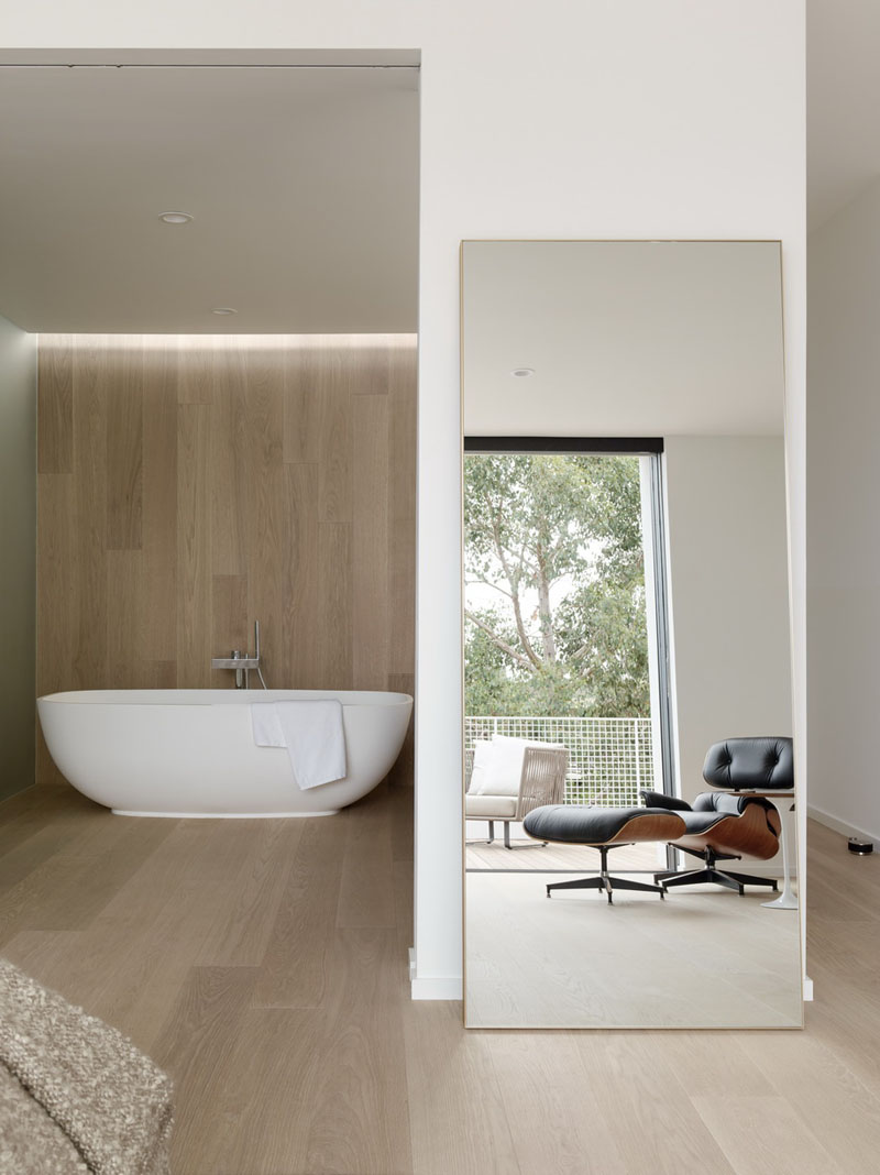 This modern bedroom and bathroom pair features a private balcony and a timber wall behind the freestanding bathtub.
