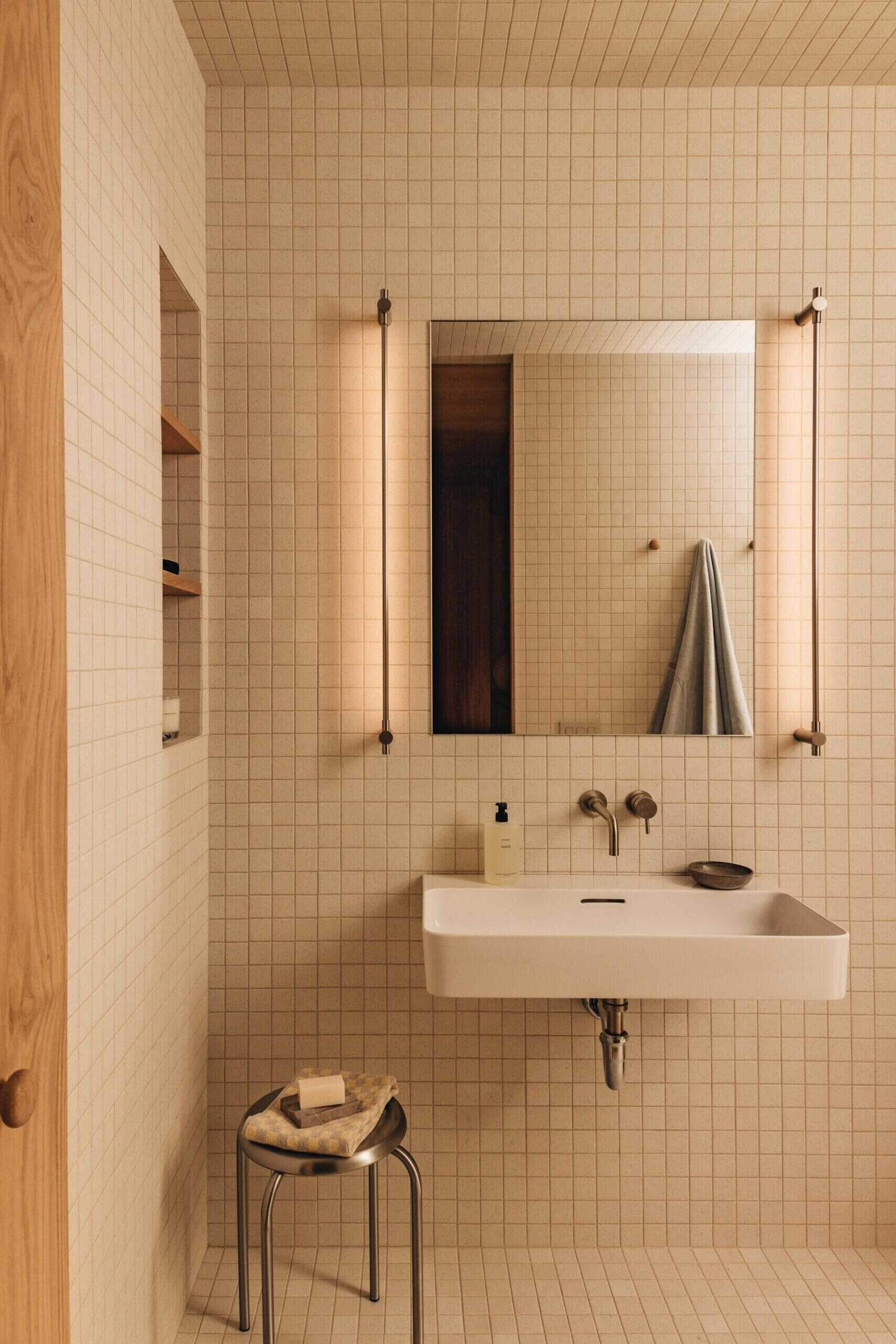 This bathroom shifts away from timber, introducing a grid of small pale tiles that brighten the room while keeping the minimalist aesthetic intact.
Wall-mounted fixtures, open shelving, slim vertical lights, and a simple rectangular basin keep the focus on proportion and material. The result feels clean, functional, and quietly elegant.