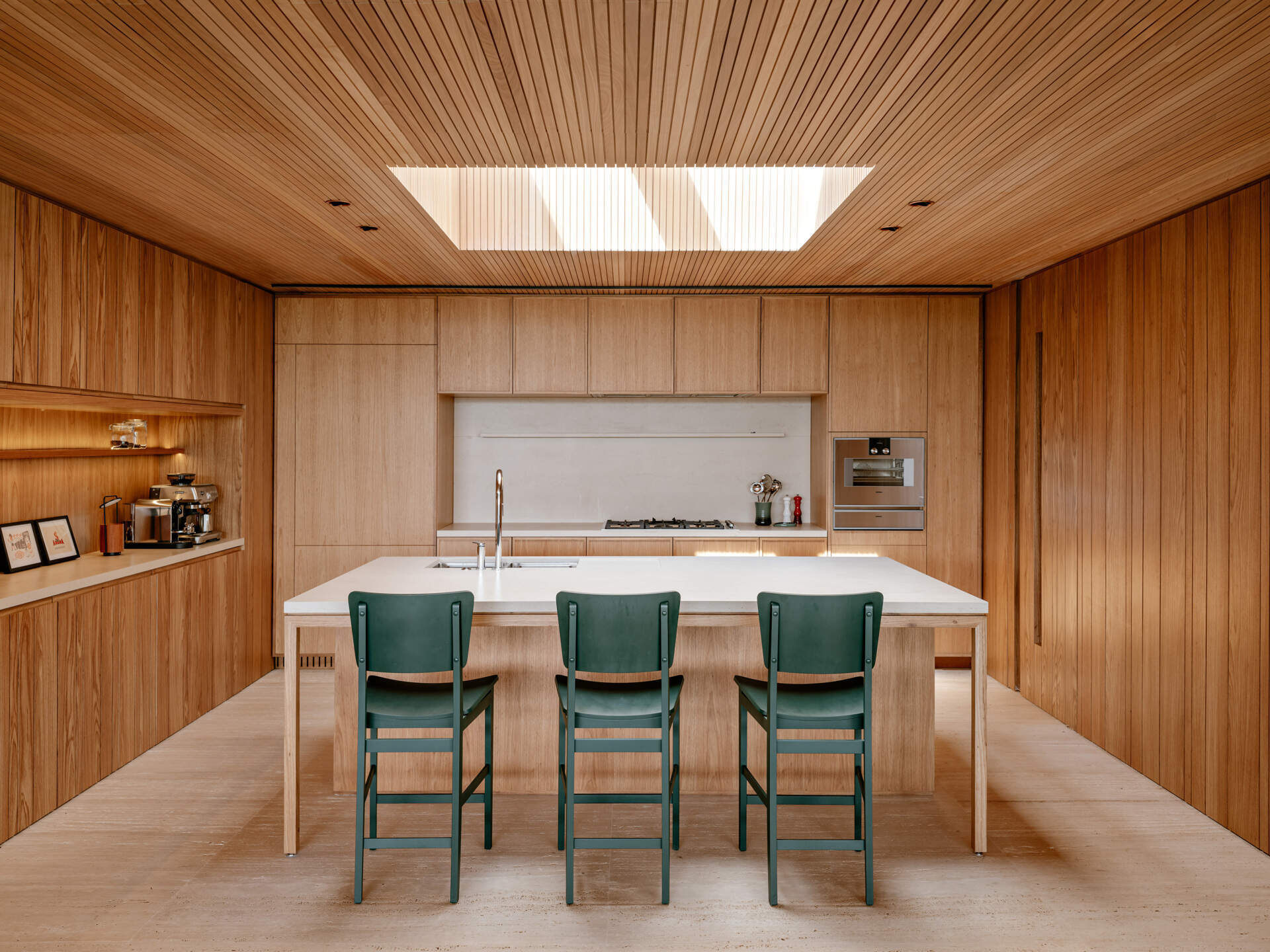 Descubra una cálida cocina de madera con tragaluces, gabinetes integrados, una gran isla blanca y taburetes verdes que agregan sutiles toques de color.