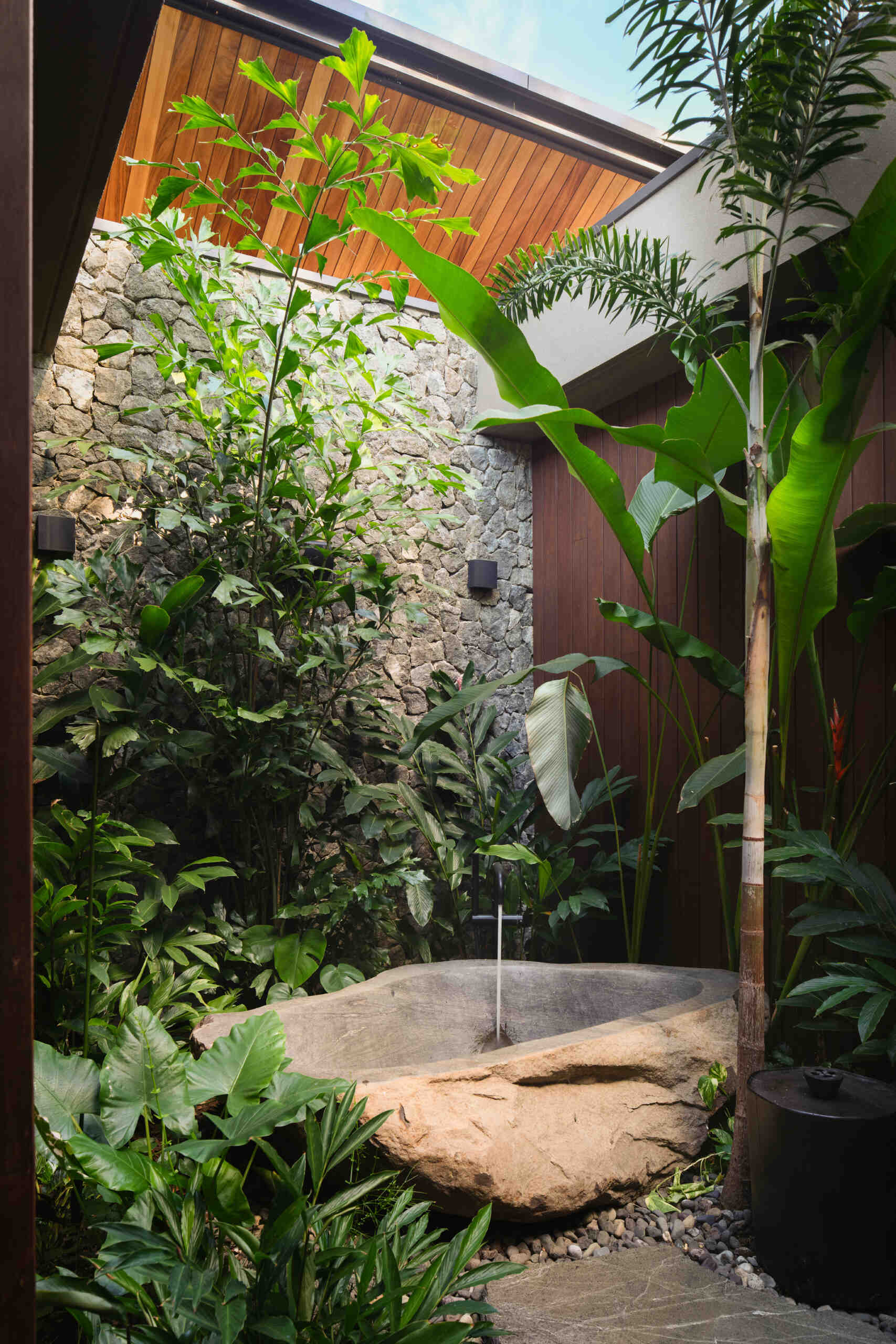 Nestled within a lush planted courtyard, the outdoor bathtub is surrounded by stone walls and tropical greenery that create a sense of privacy. Sunlight filters through the opening above, while nearby a timber-lined ceiling introduces warmth to the sheltered bathing area.