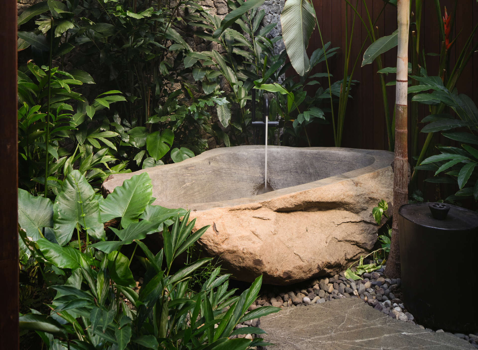 Nestled within a lush planted courtyard, the outdoor bathtub is surrounded by stone walls and tropical greenery that create a sense of privacy. Sunlight filters through the opening above, while nearby a timber-lined ceiling introduces warmth to the sheltered bathing area.
