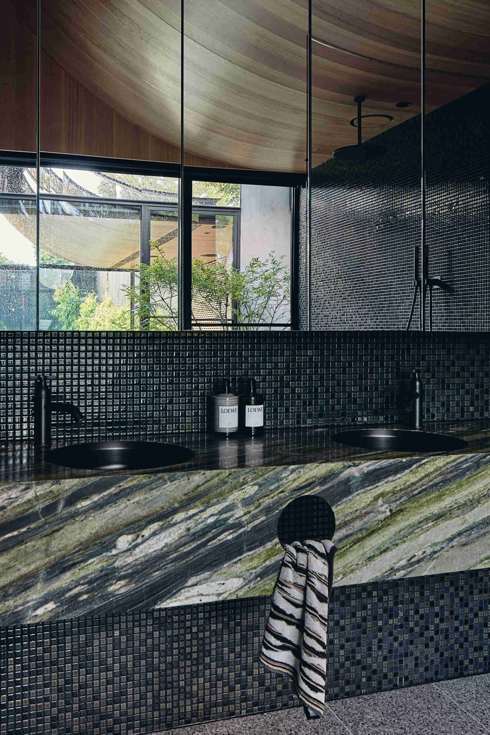 A primary bathroom combines warm wood paneling, dark mosaic tiles and richly veined green stone, with expansive garden views, a sculptural freestanding bath and adjoining dressing area.