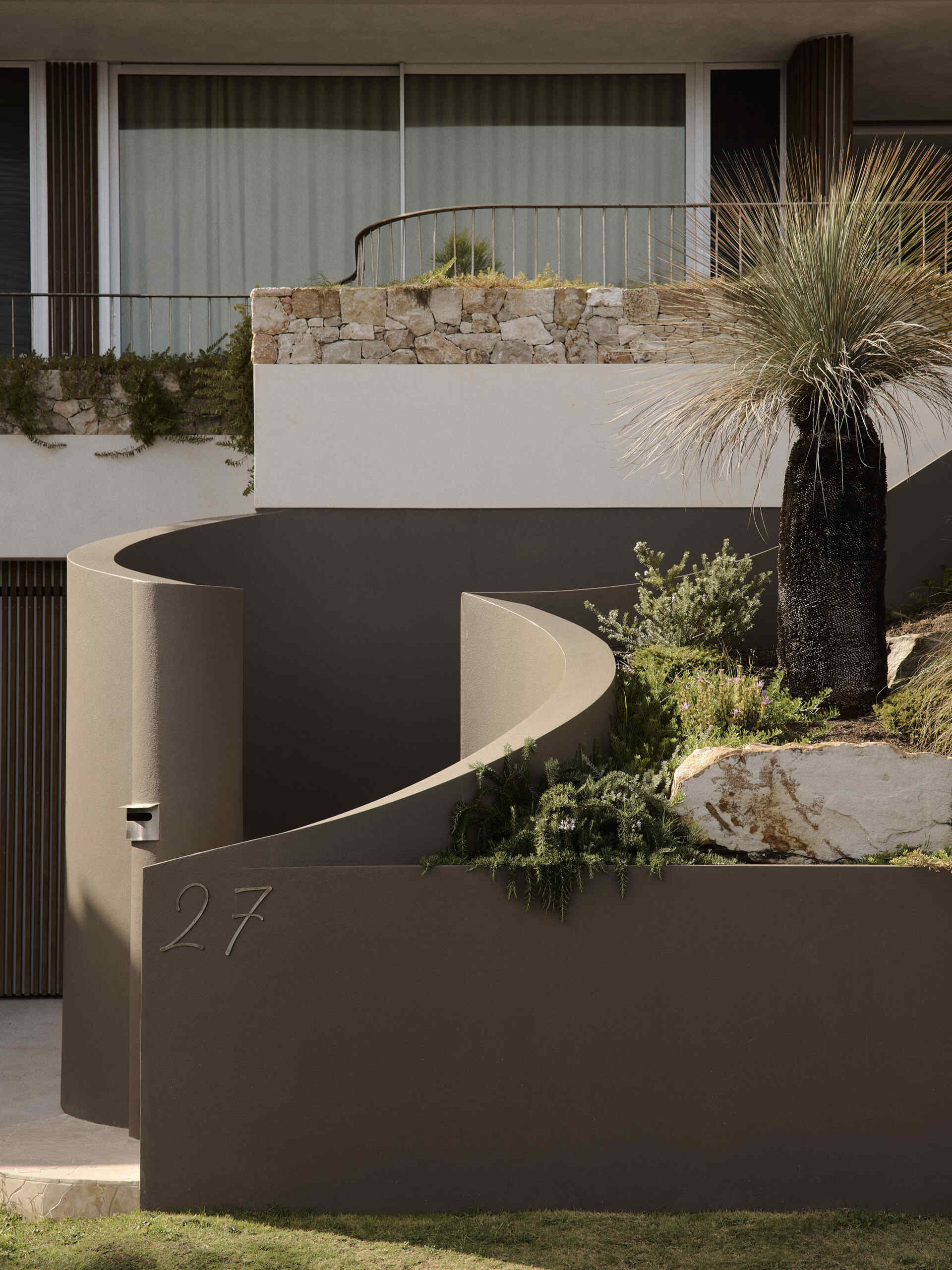 Curved taupe stucco walls, pale stone steps and lush planting create a sculptural entrance to this Byron Bay home, softened by a mature grass tree and touches of purple flowers.