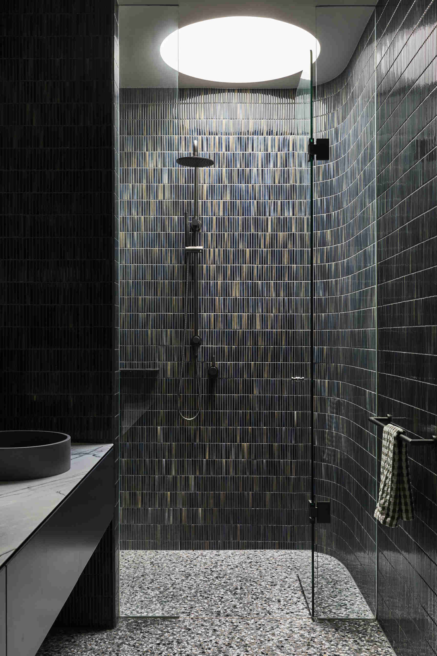 Discover a compact bathroom at Sasso Nero with deep green vertical tiles, a curved glass shower, pale marble vanity, terrazzo flooring and matte black fixtures beneath a large circular ceiling light.