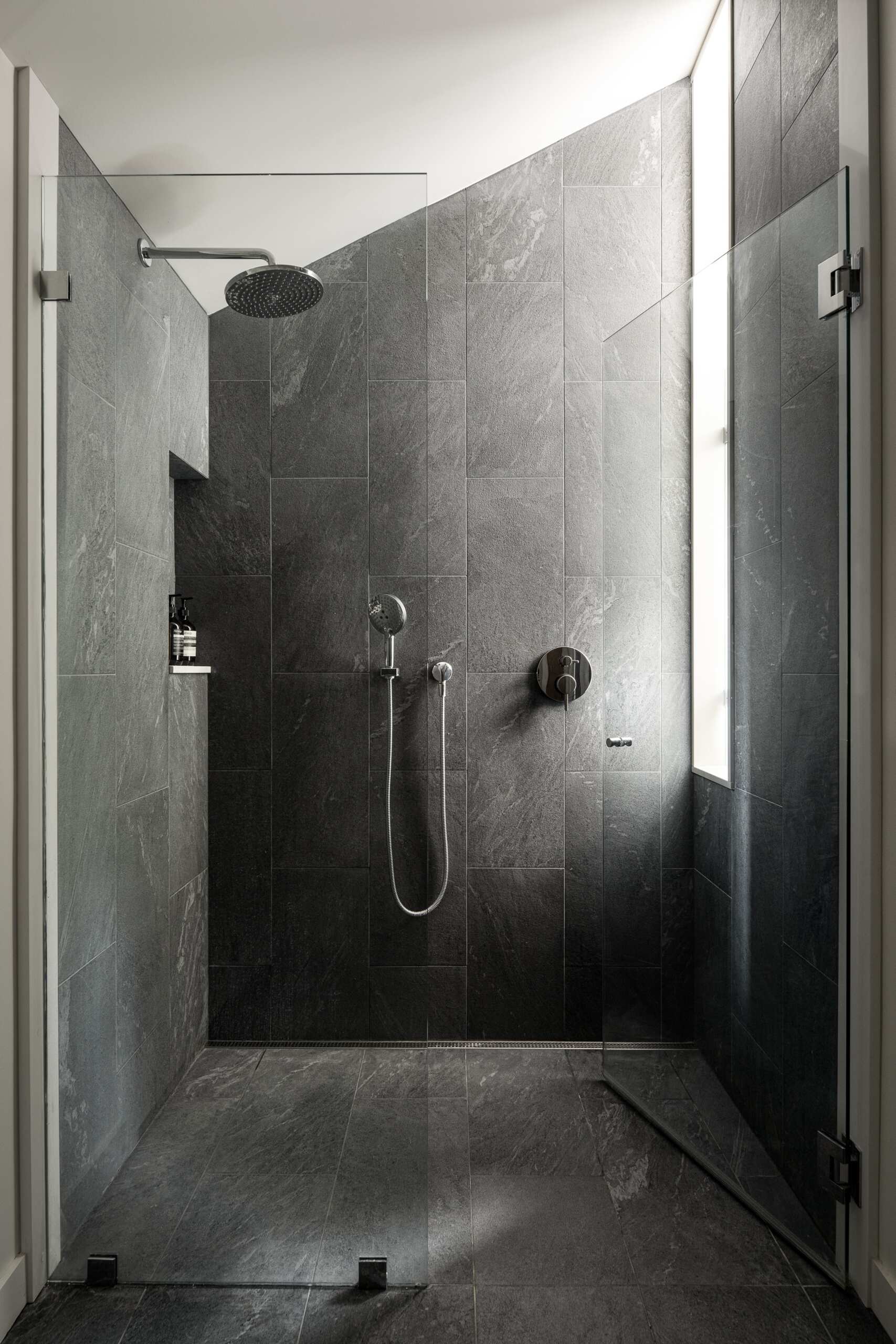 The guest bathroom takes a darker approach, with grey slate-look tiles covering the shower walls and floor. The large-format tiles give the space a consistent material palette, while the varied surface markings add texture across the walls.