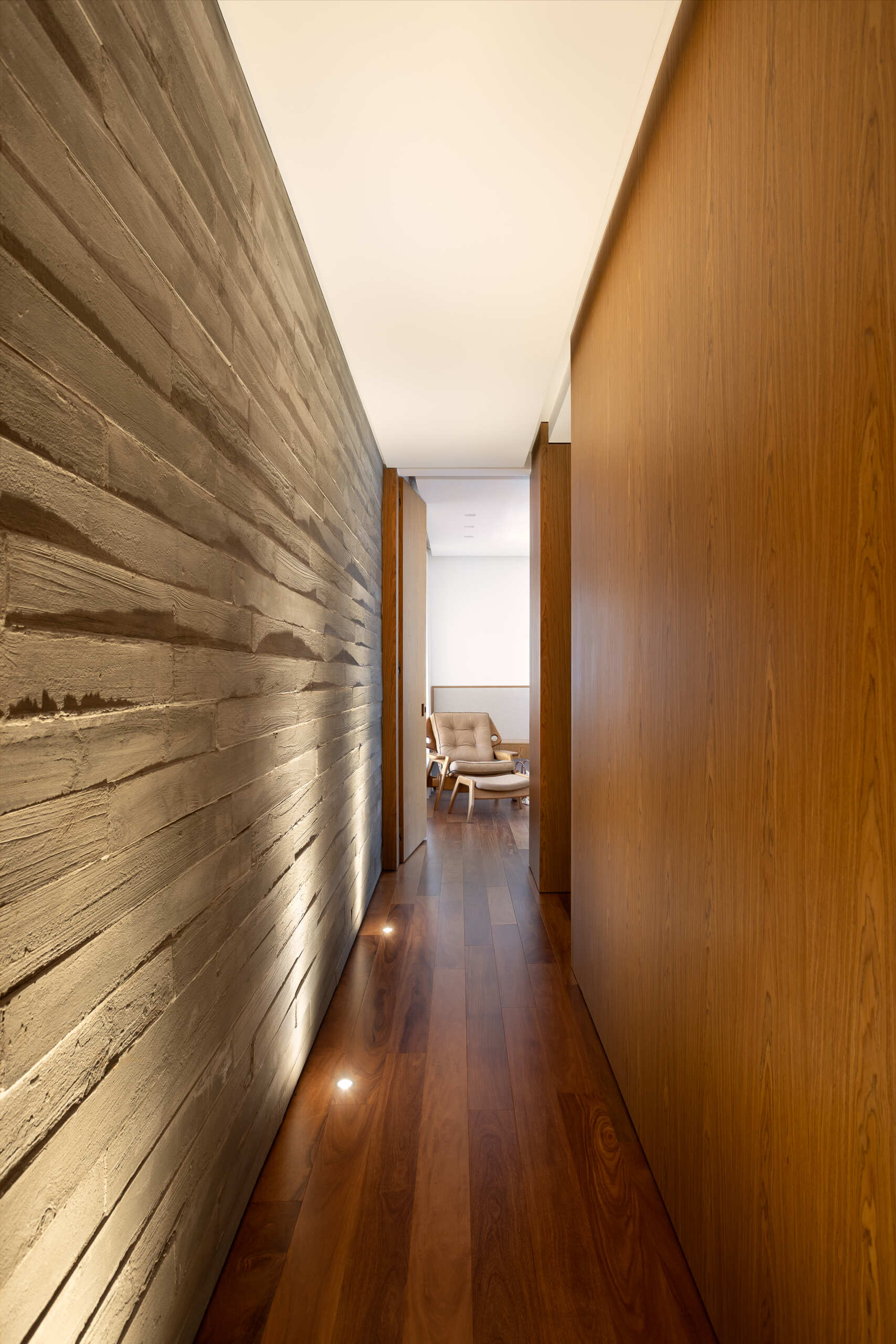 The primary bedroom at Casa da Mata combines soft neutrals, warm wood, sheer curtains and a caramel leather lounge chair, with views toward the pool and tropical greenery outside.