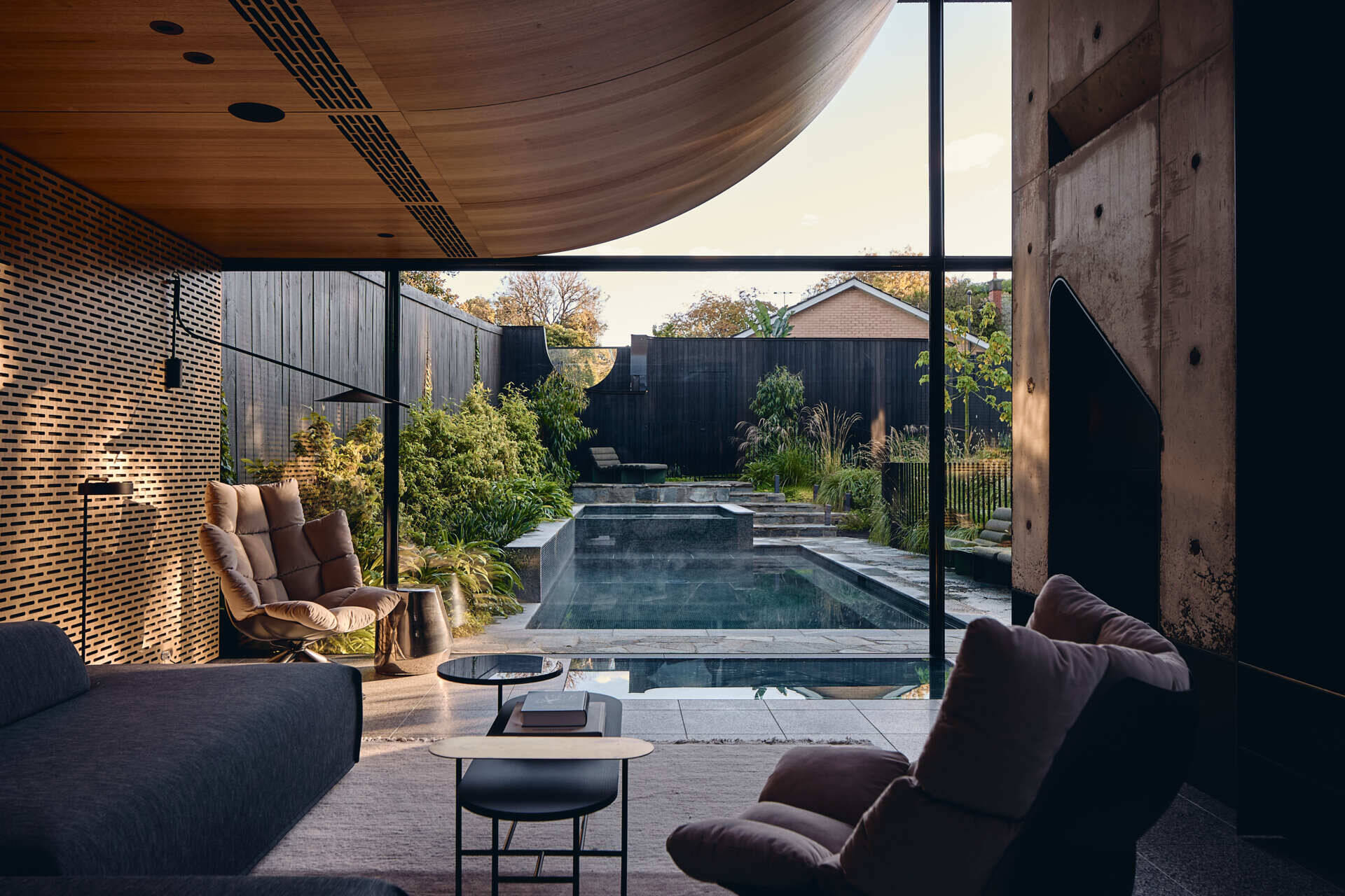A timber-lined living room at Babylonia overlooks the pool and garden, with curved ceilings and framed views toward the greenery surrounding this Melbourne home.