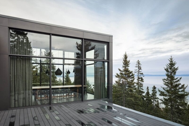 A glass-railed deck extends from the social spaces of this Québec home, allowing the river and surrounding landscape to remain visible from inside and outside.