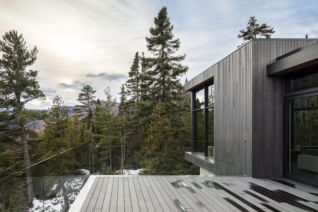 A glass-railed deck extends from the social spaces of this Québec home, allowing the river and surrounding landscape to remain visible from inside and outside.