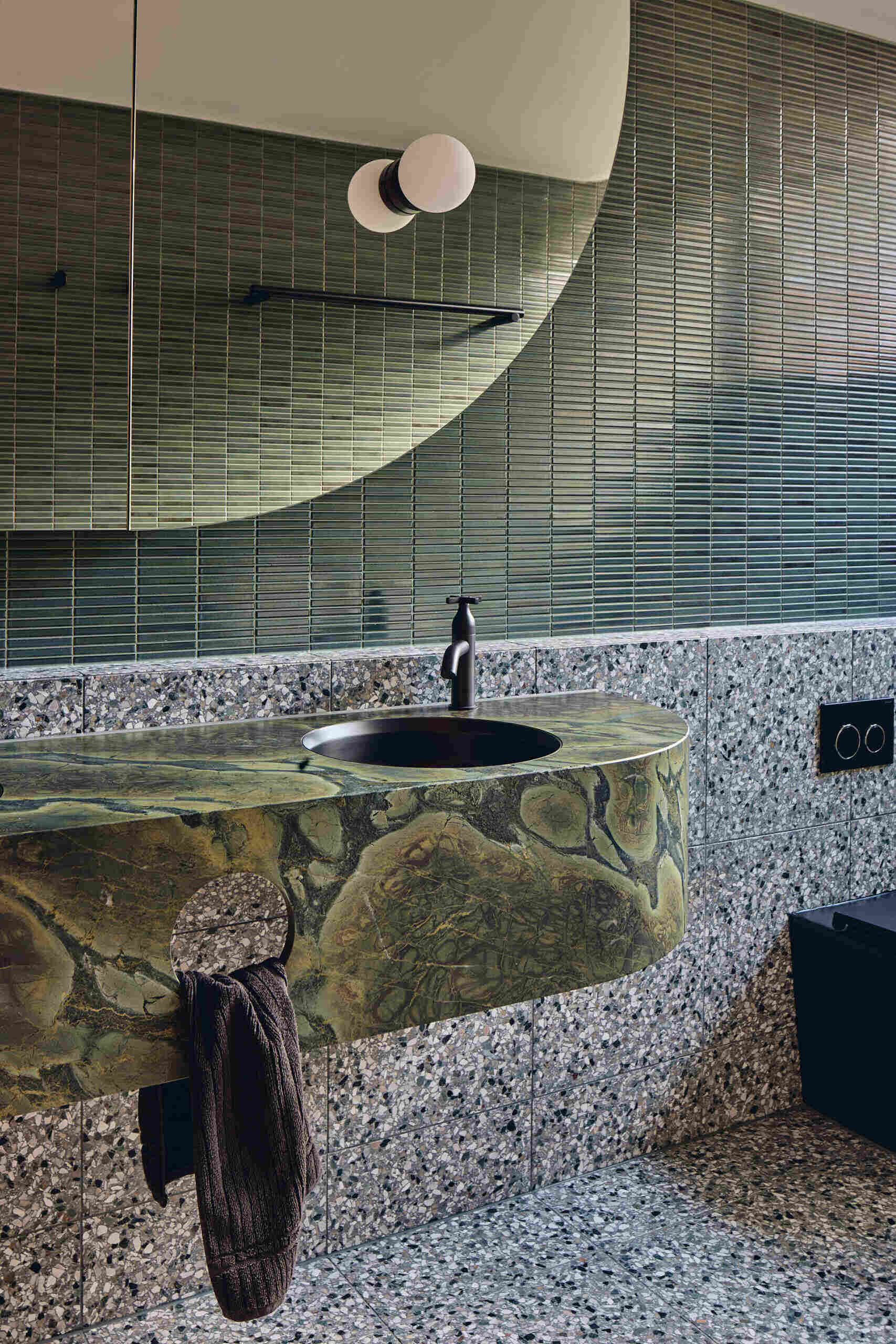 Deep green tiles, speckled terrazzo and richly veined stone give this Babylonia guest bathroom a layered material palette, finished with a curved mirror and matte black fixtures.