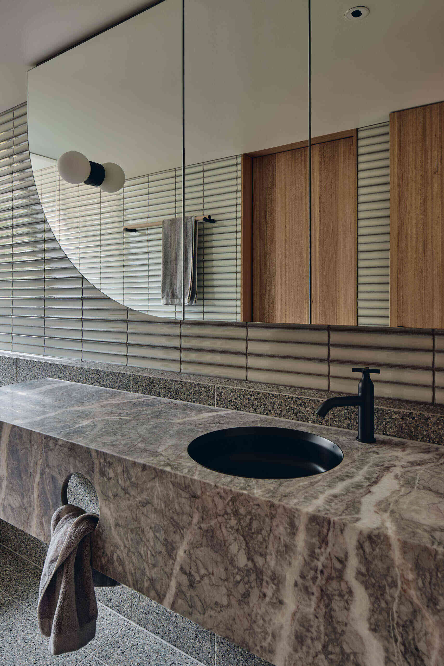 A richly veined stone vanity, pale horizontal tiles, warm wood and speckled terrazzo come together in this guest bathroom at Babylonia by FMD Architects.