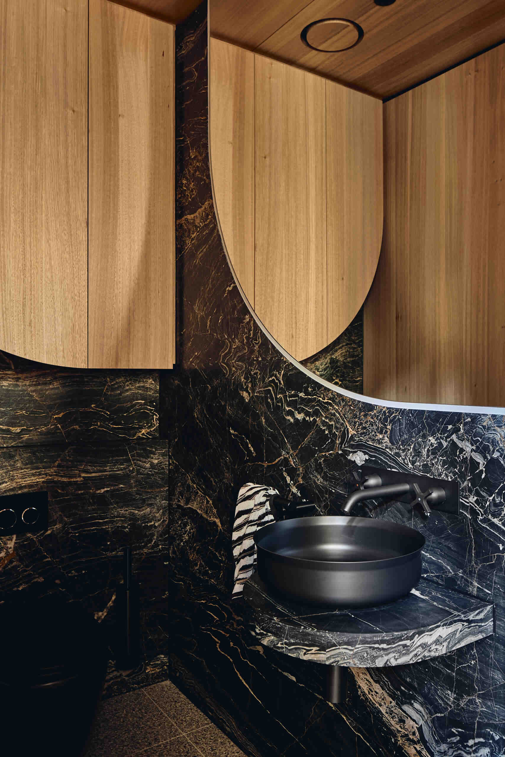 Black stone and warm timber shape this compact guest bathroom at Babylonia, with a round black vessel sink and curved mirror adding softer forms to the space.