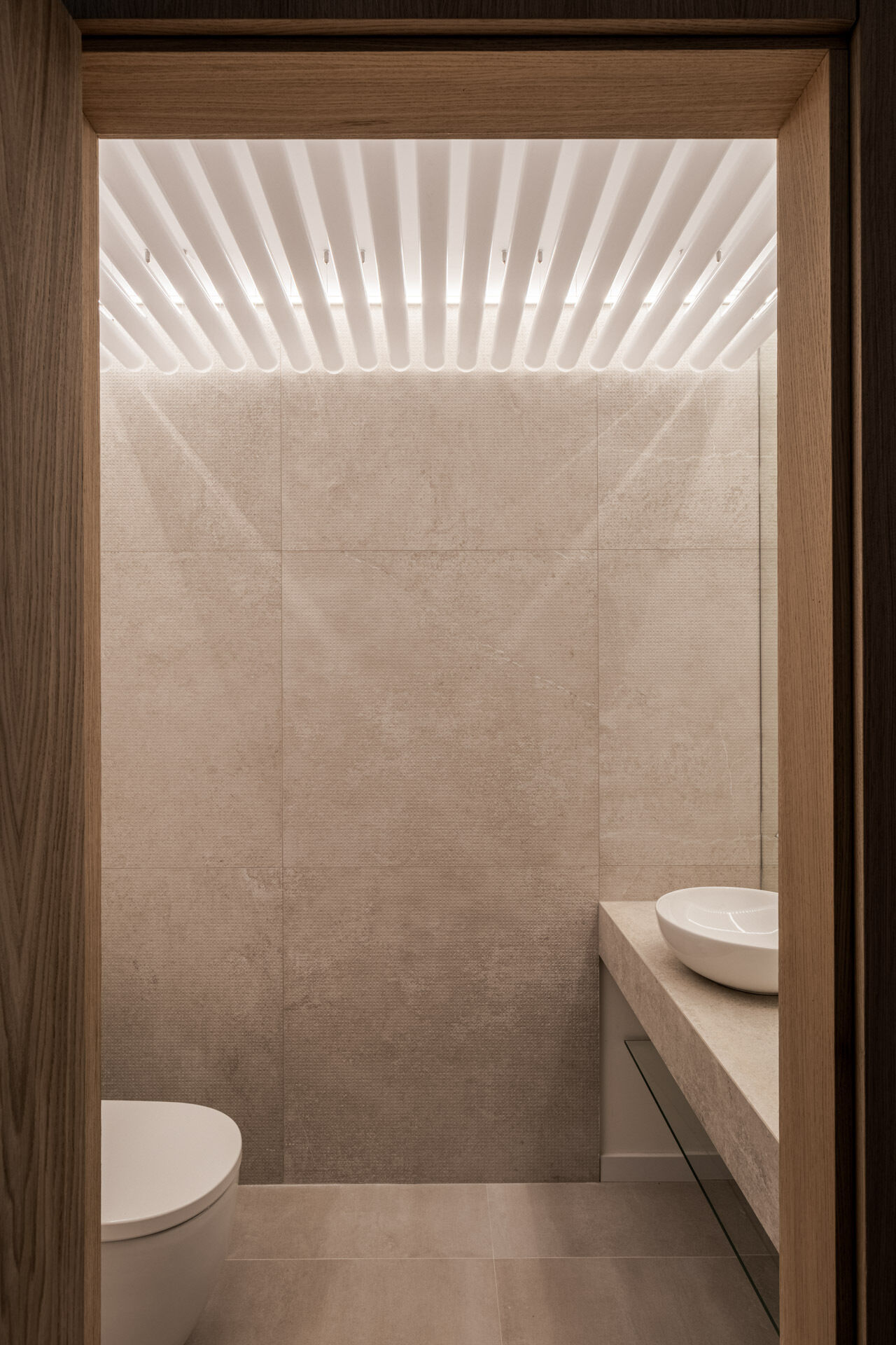 A sculptural powder room inside the central timber volume features curved backlit slats, sand-toned textured walls and an organic freestanding basin.