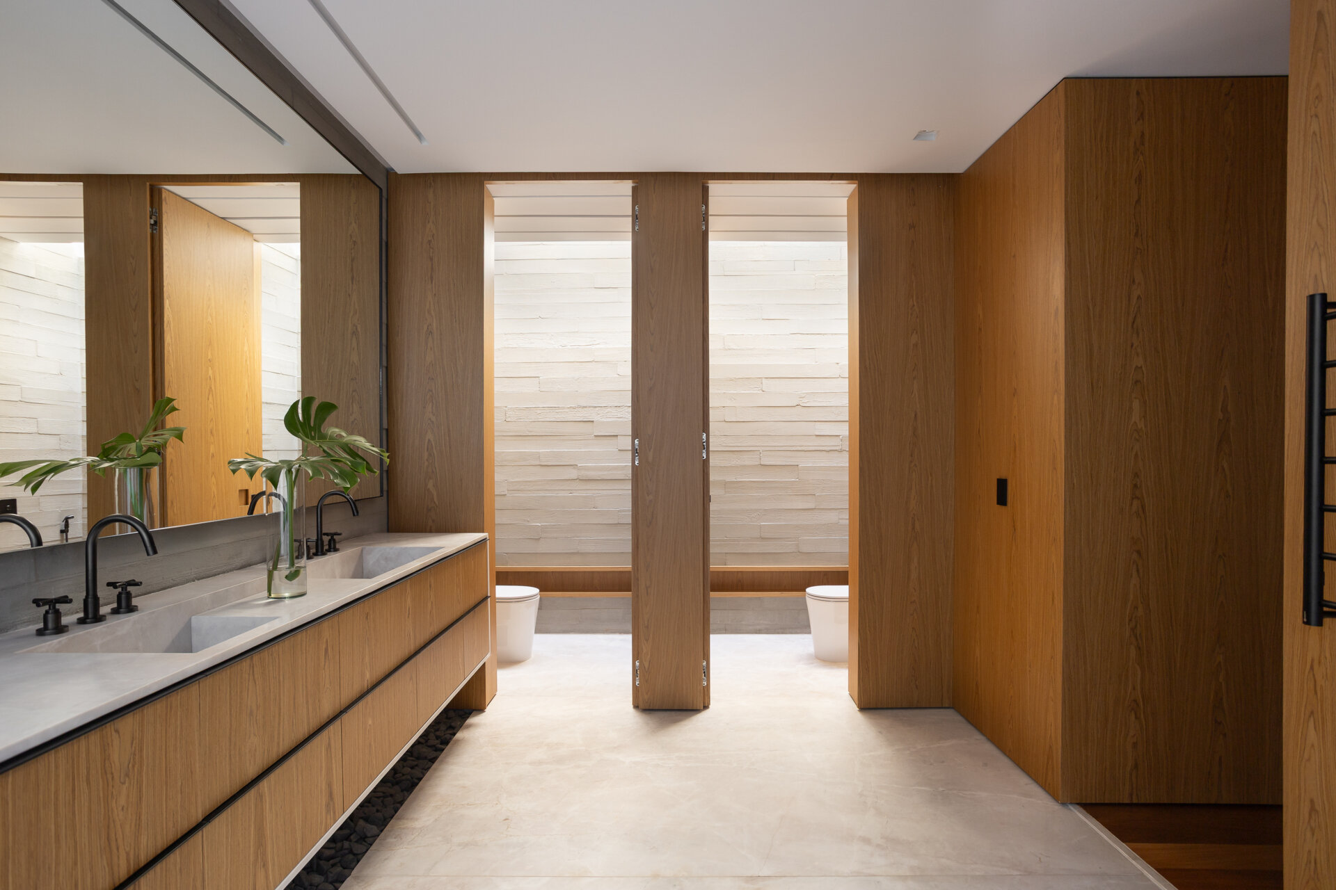 The primary bathroom at Casa da Mata combines warm wood cabinetry, pale stone and black fixtures with a long double vanity, full-height mirror and private planted courtyard.