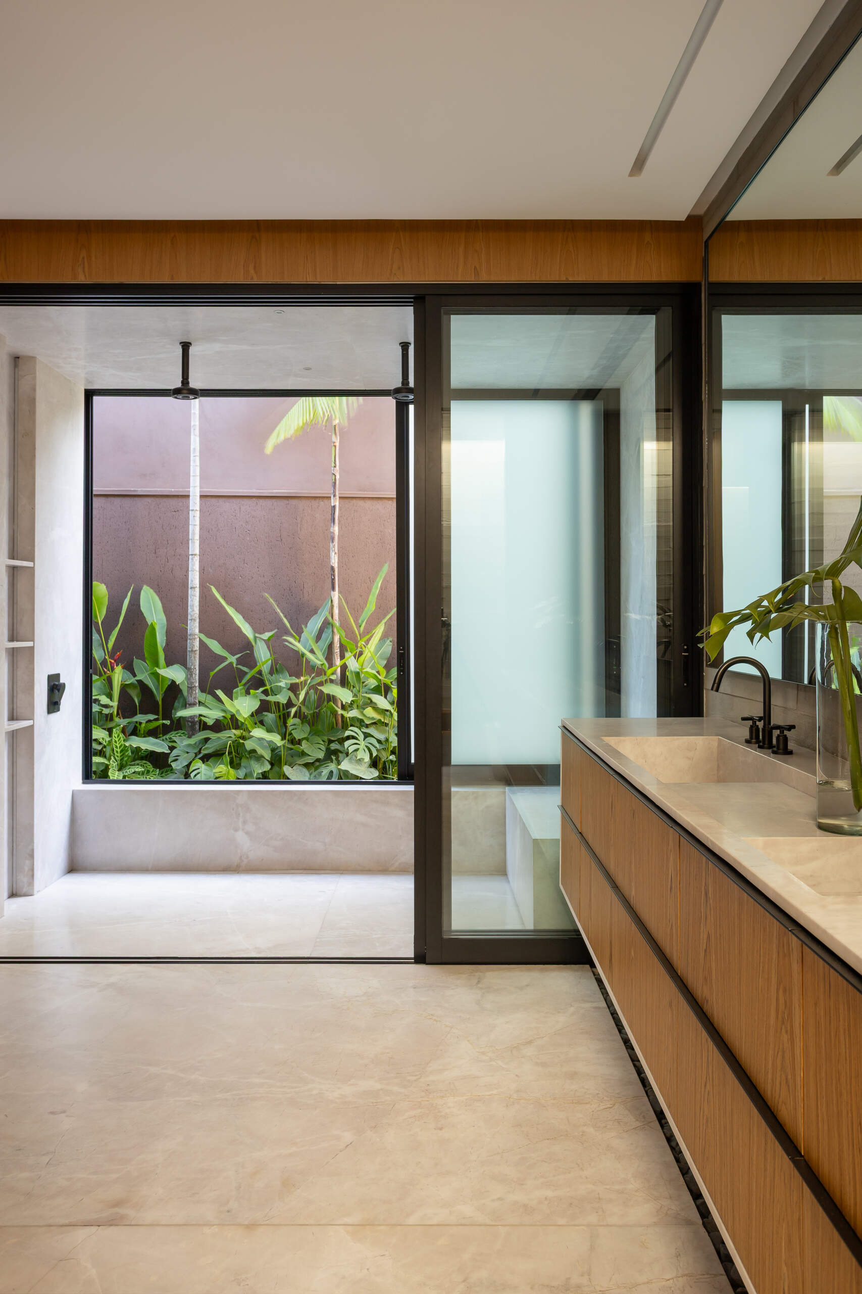 The primary bathroom at Casa da Mata combines warm wood cabinetry, pale stone and black fixtures with a long double vanity, full-height mirror and private planted courtyard.