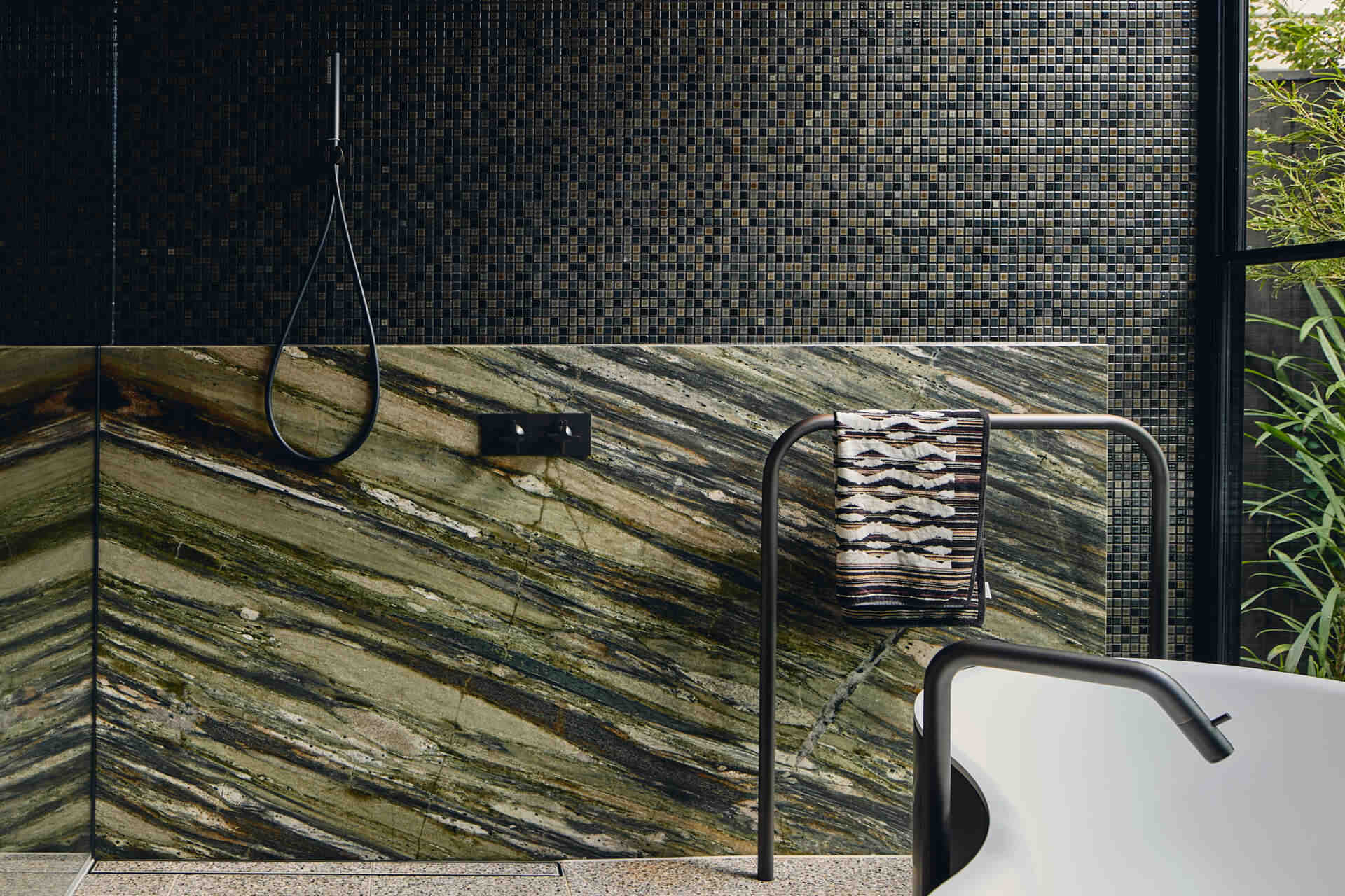 A primary bathroom combines warm wood paneling, dark mosaic tiles and richly veined green stone, with expansive garden views, a sculptural freestanding bath and adjoining dressing area.