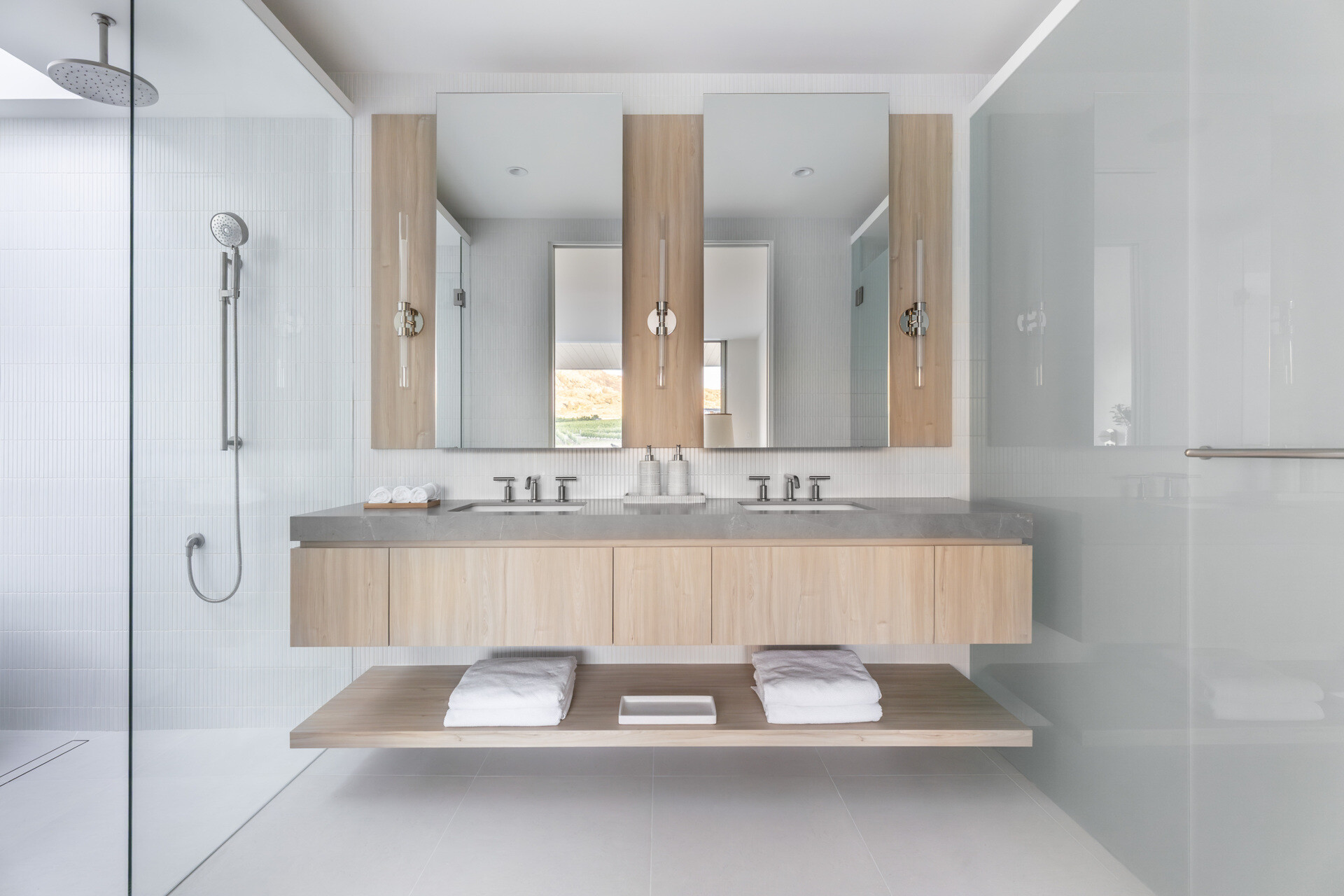 Take a look inside this bright, spa-like bathroom with pale wood cabinetry, gray stone counters, white tile, a floating double vanity, freestanding tub, and skylights over the shower and bathing area.