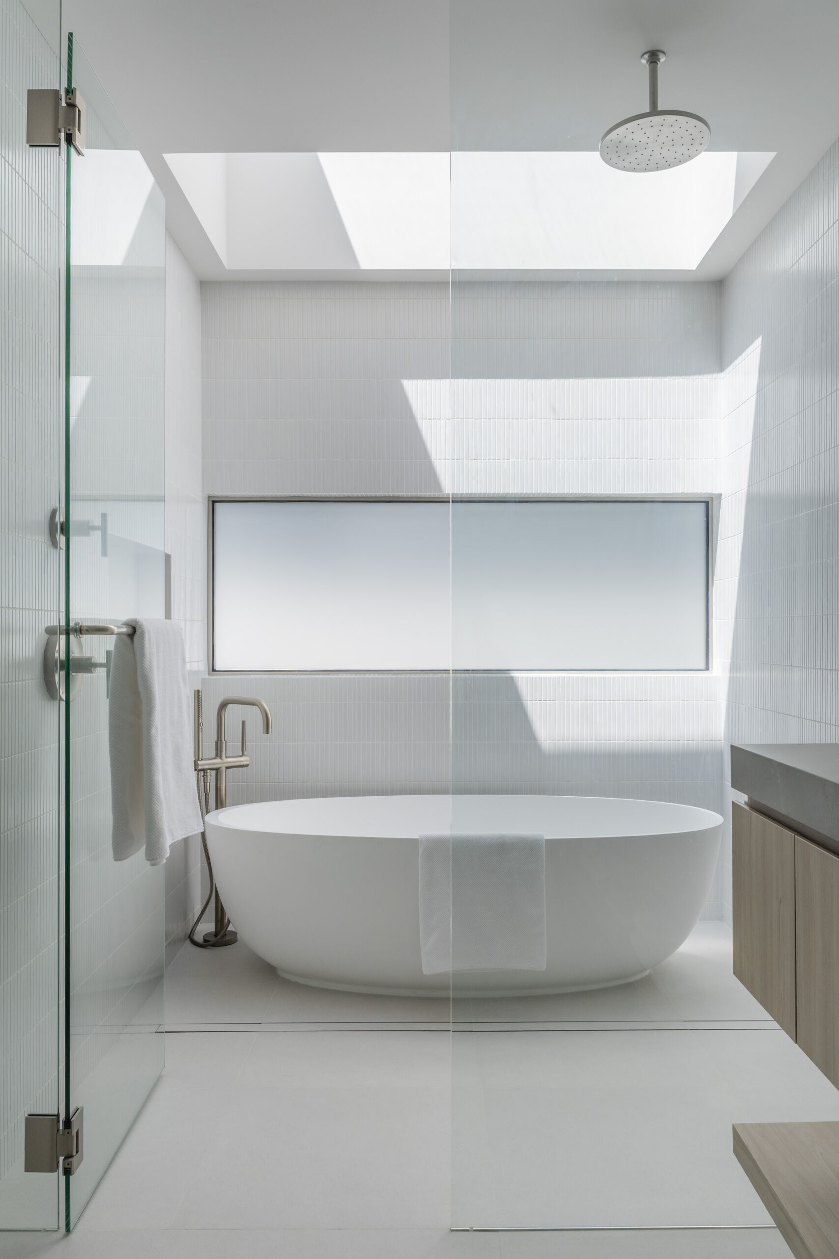 Take a look inside this bright, spa-like bathroom with pale wood cabinetry, gray stone counters, white tile, a floating double vanity, freestanding tub, and skylights over the shower and bathing area.