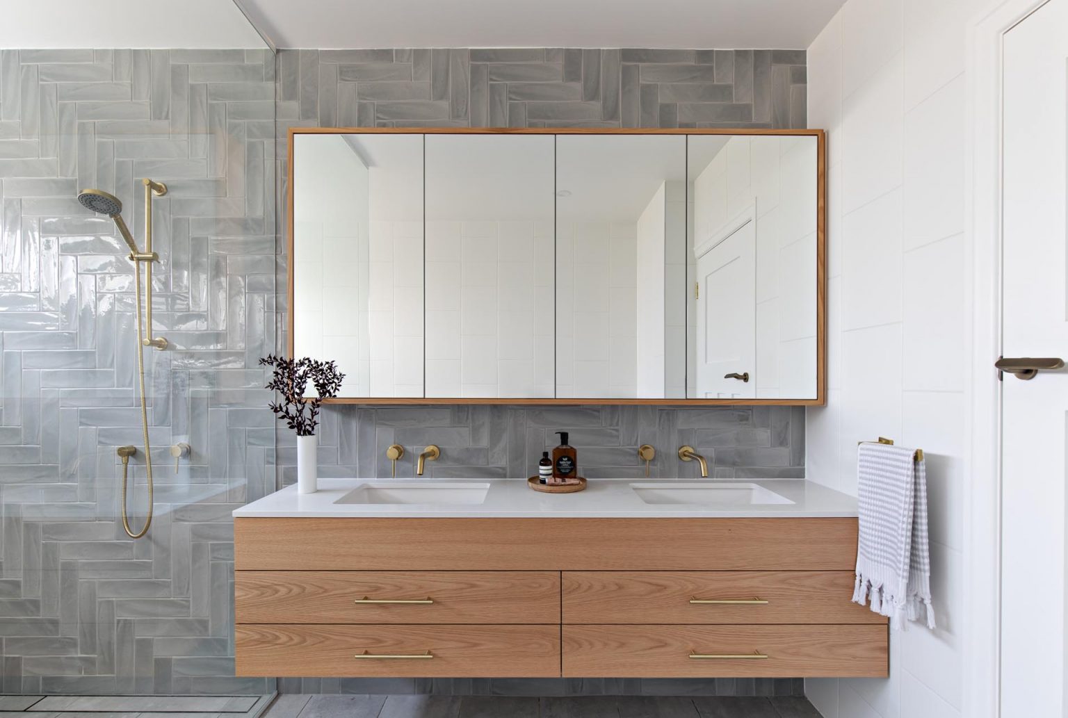 Explore a classic contemporary bathroom with grey herringbone tiles, a floating wood vanity, white countertop, freestanding bathtub and glass shower screen.