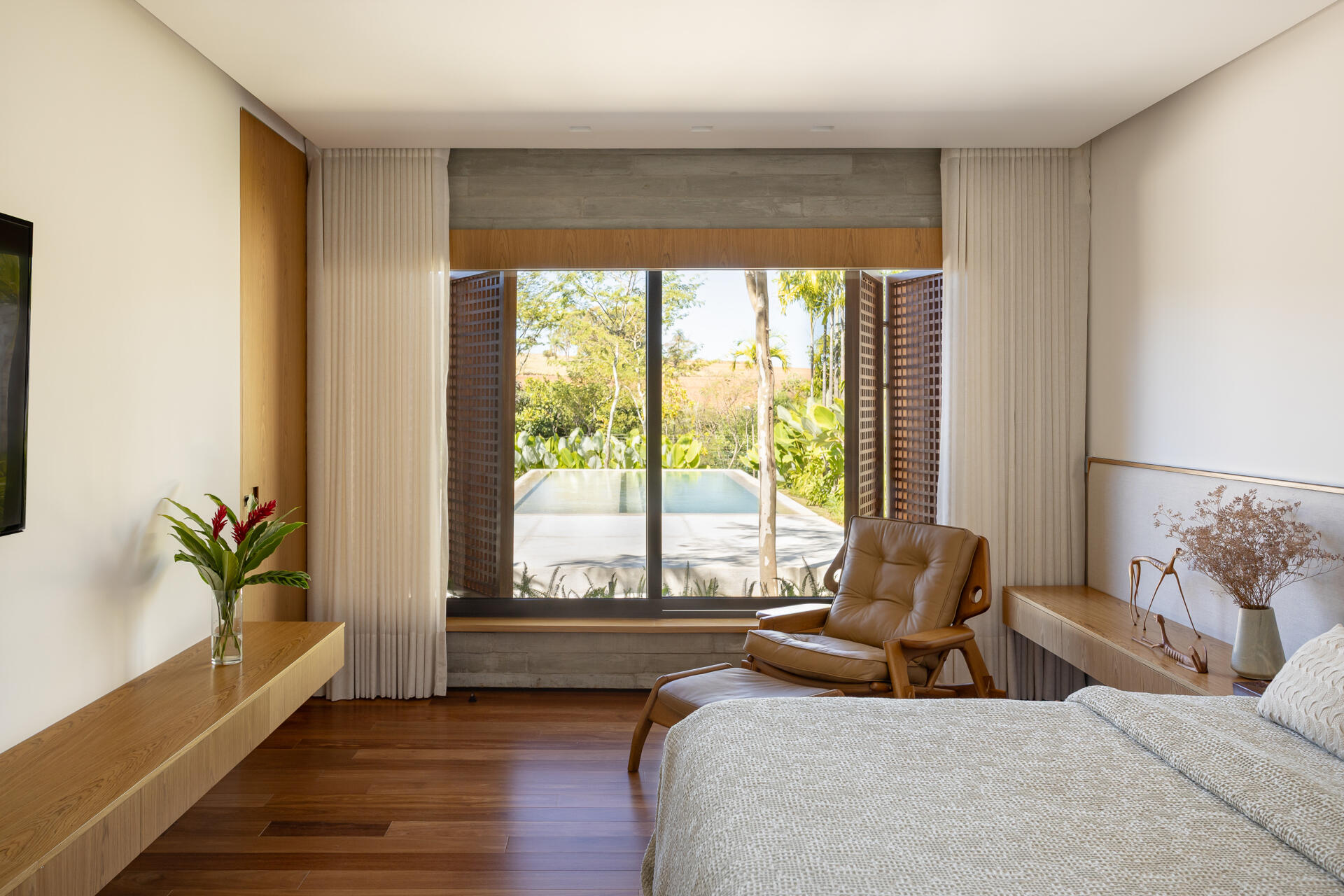 The primary bedroom at Casa da Mata combines soft neutrals, warm wood, sheer curtains and a caramel leather lounge chair, with views toward the pool and tropical greenery outside.