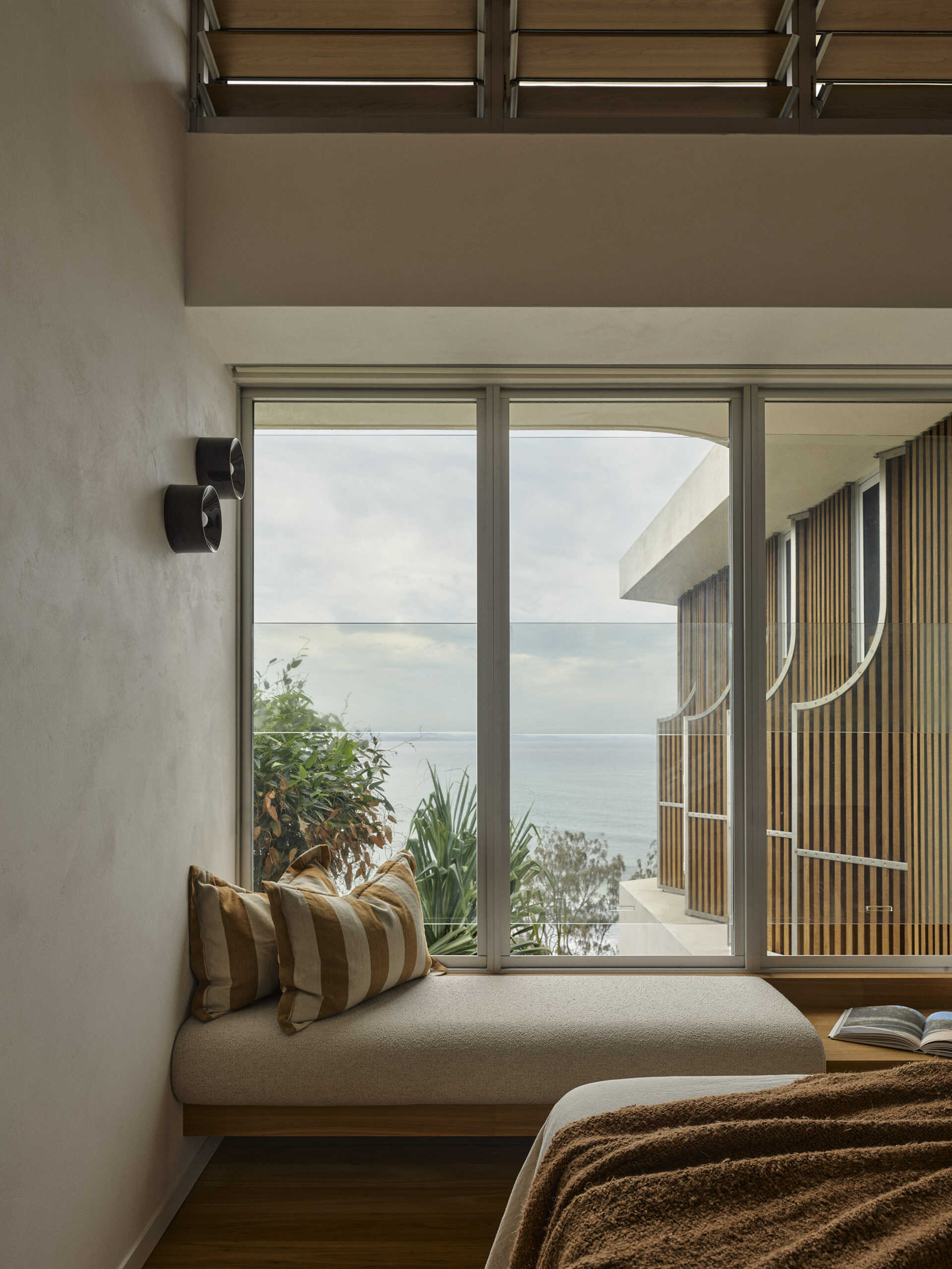 A cosy guest bedroom in this Byron Bay beach house features a built-in window bench, warm natural finishes and generous glazing that makes the most of the surrounding coastal views.