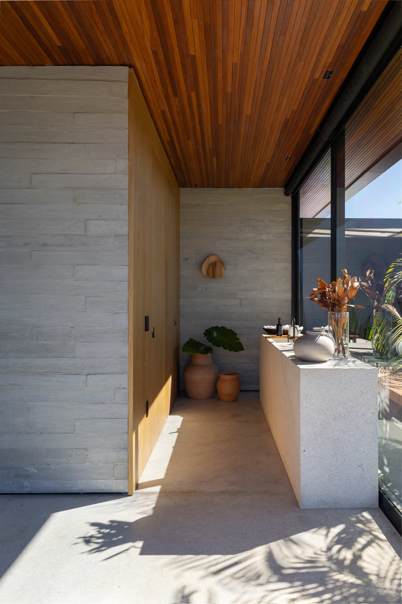 A compact powder room behind the kitchen at Casa da Mata combines warm wood, pale concrete, a long stone vanity, full-height glass and natural decorative details.