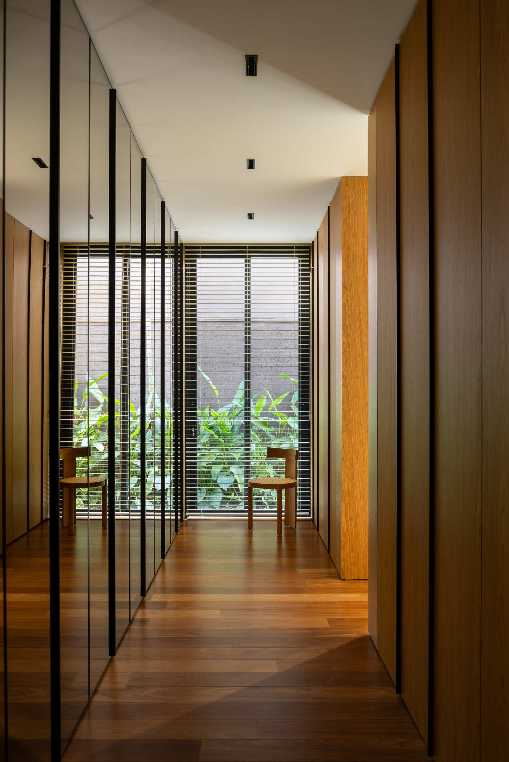 Warm wood floors, full-height cabinetry and mirrored doors define the walk-in closet in Casa da Mata's primary suite, with black-framed glazing and greenery outside adding contrast.