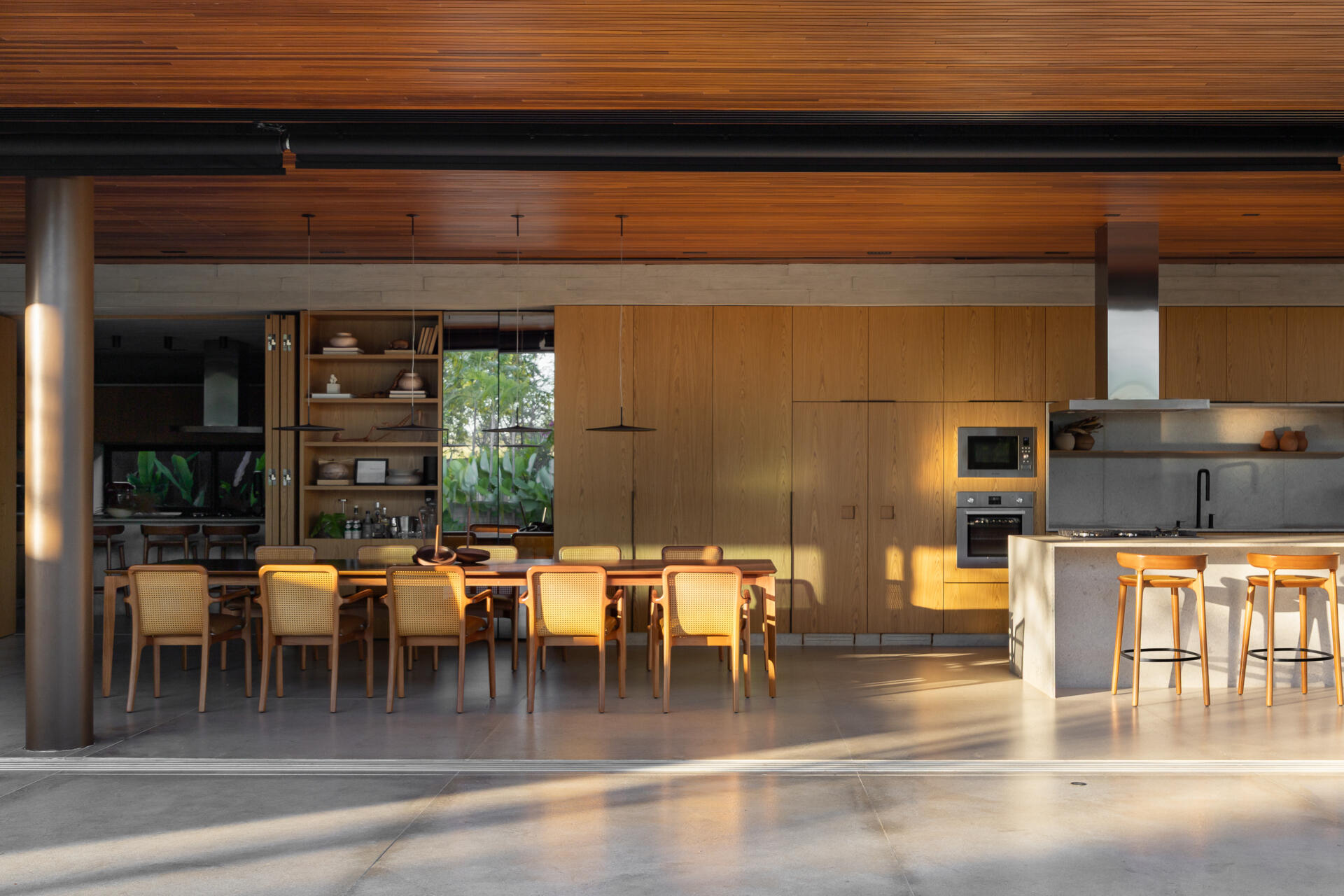 This open-plan kitchen and dining area at Casa da Mata combines natural wood cabinetry, stone surfaces, concrete floors, woven dining chairs and a rich wood-lined ceiling, with wide openings connecting to the tropical garden.