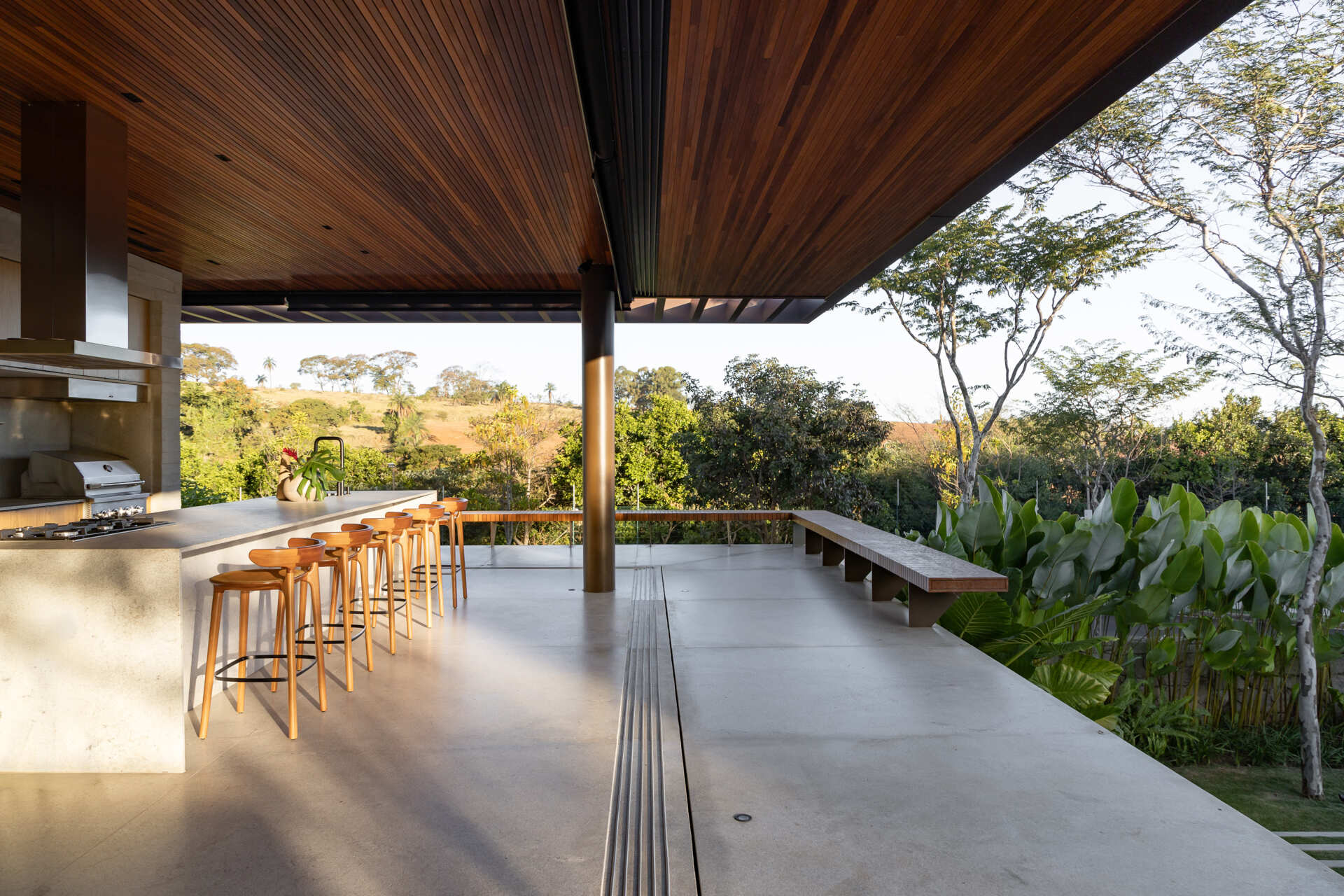 This open-plan kitchen and dining area at Casa da Mata combines natural wood cabinetry, stone surfaces, concrete floors, woven dining chairs and a rich wood-lined ceiling, with wide openings connecting to the tropical garden.