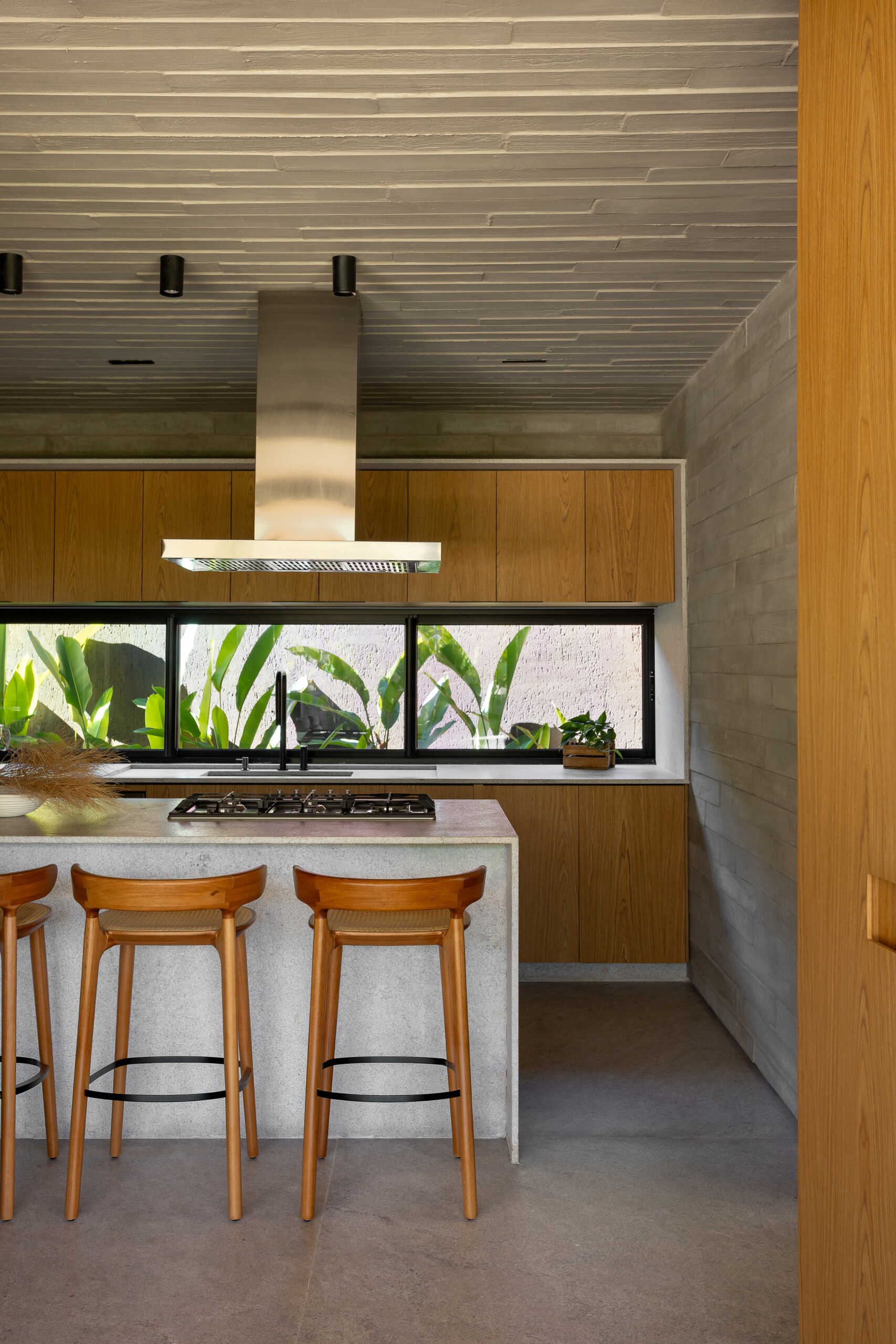 The butler's kitchen at Casa da Mata pairs warm wood cabinetry with cool gray concrete walls, stone surfaces and a textured ceiling, while a wide horizontal window frames greenery outside.