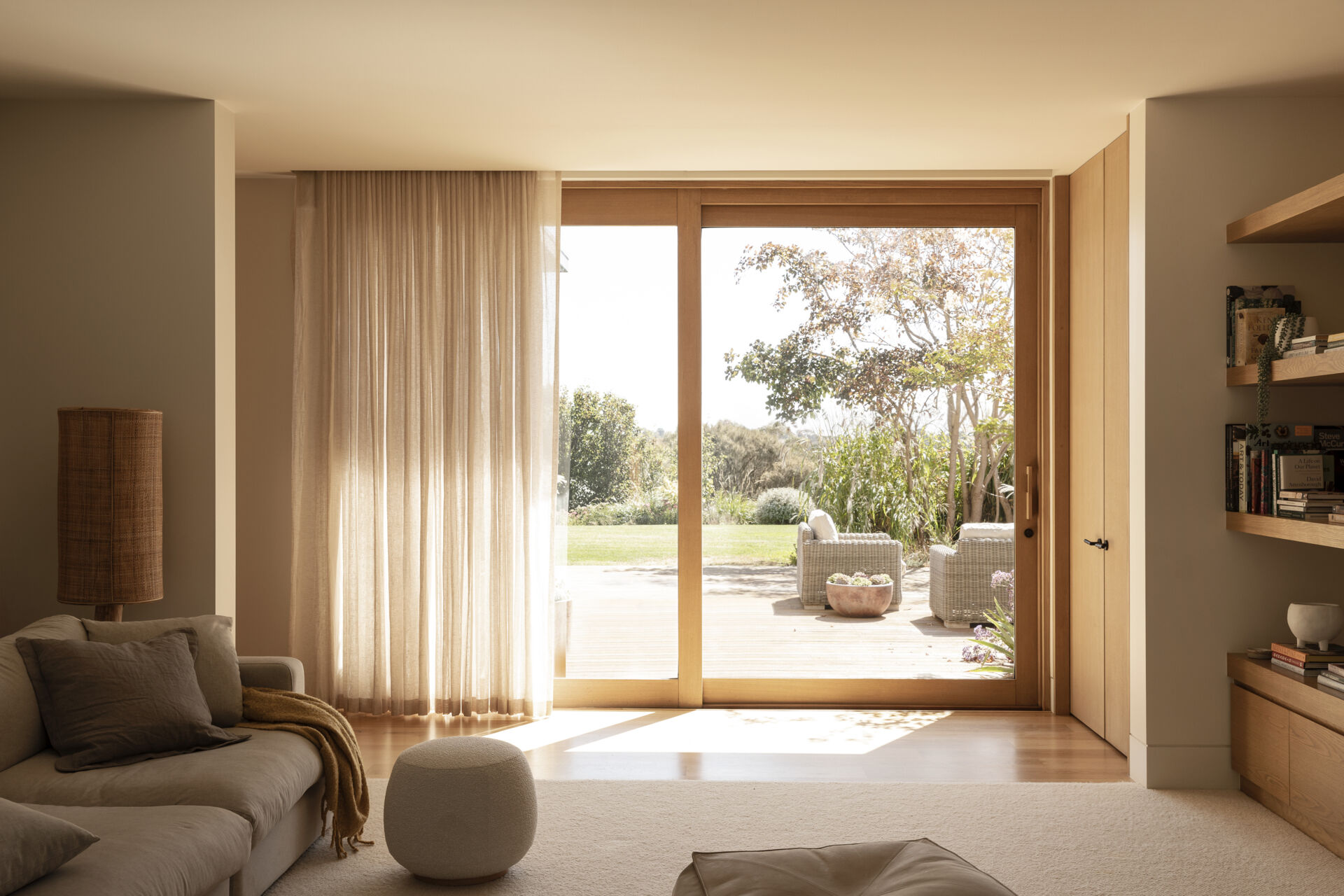 Soft linen curtains filter the sunlight in the family room, while pale wood floors, built-in shelving and warm wood-framed doors add subtle warmth. A generously cushioned sectional layered with pillows and a textured throw makes the room feel especially inviting.