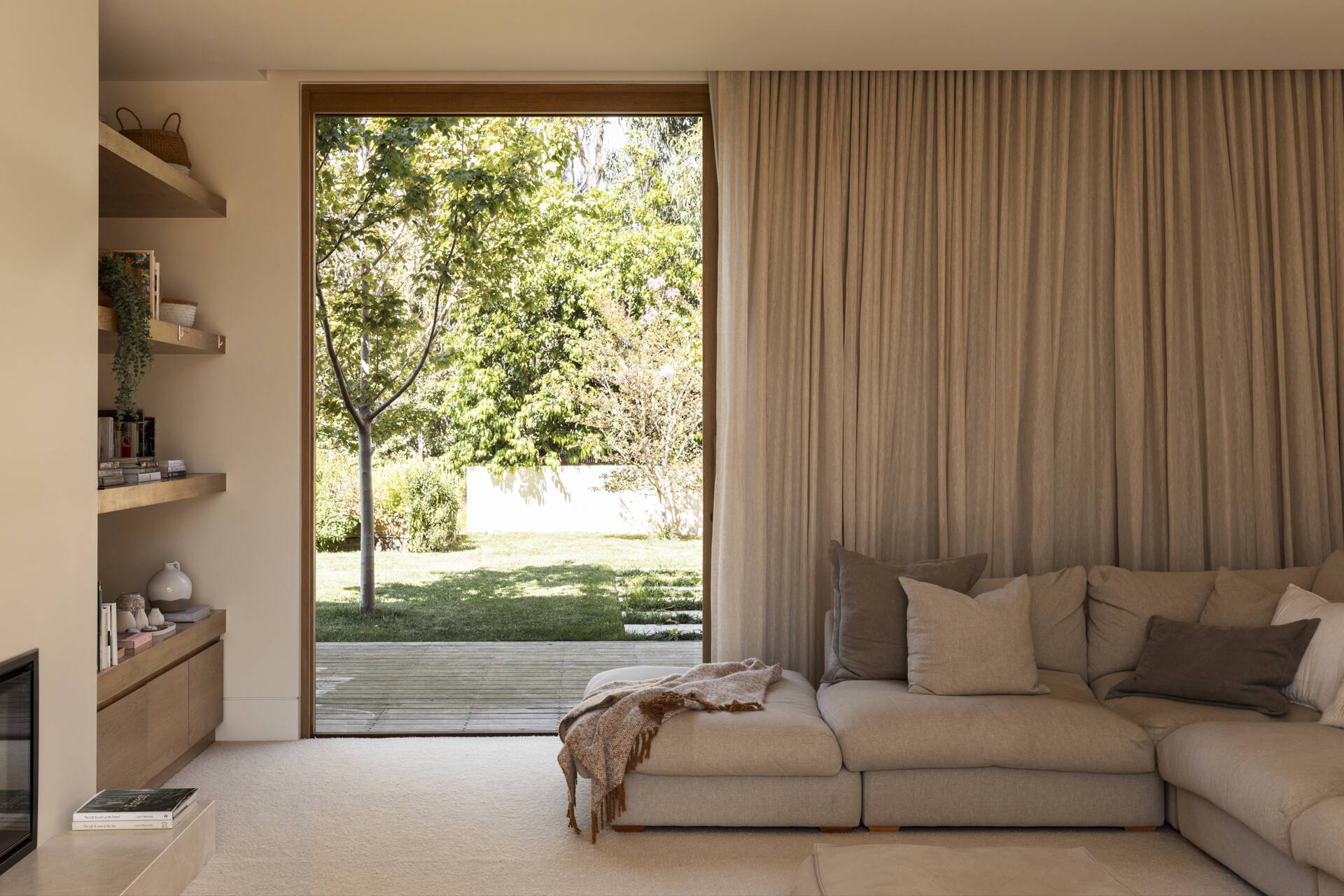 Soft linen curtains filter the sunlight in the family room, while pale wood floors, built-in shelving and warm wood-framed doors add subtle warmth. A generously cushioned sectional layered with pillows and a textured throw makes the room feel especially inviting.