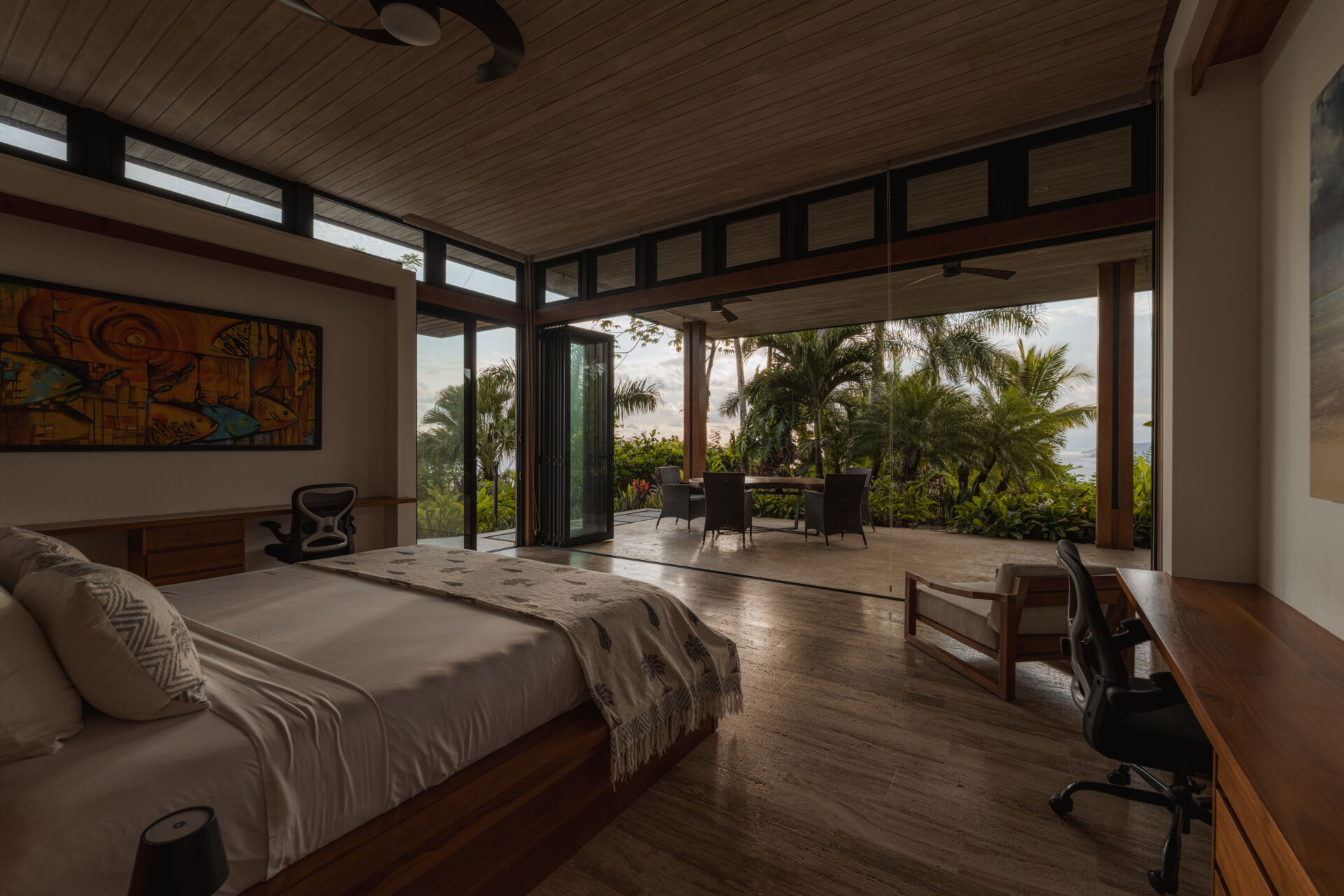 Warm wood, woven textures, colorful artwork and expansive glass doors define the tropical bedroom at Tierra y Mar House, opening toward palms and ocean views.