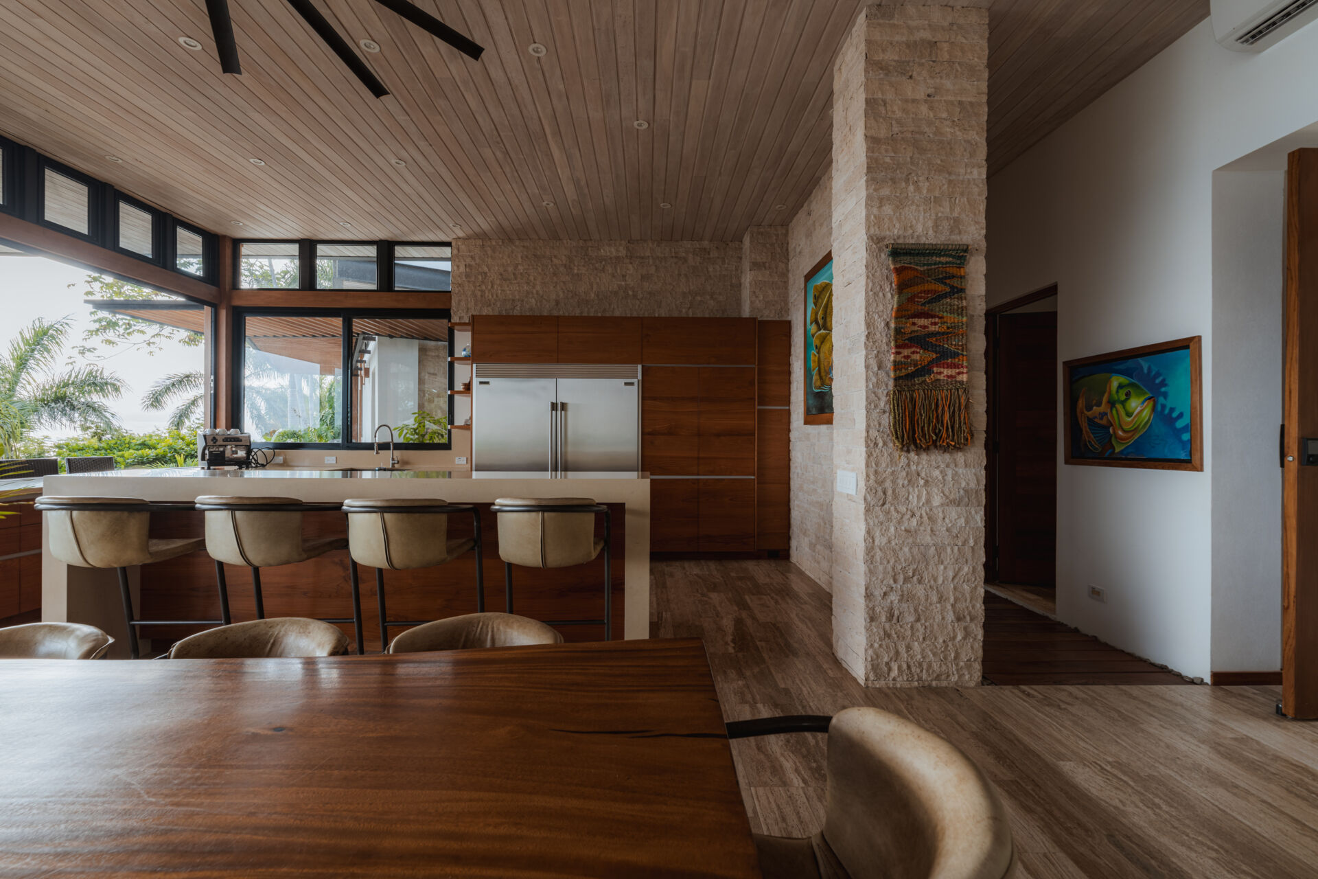 Warm wood cabinetry, pale stone surfaces and a textured stone column define the open kitchen at Tierra y Mar House, with greenery and natural light flowing in through wide windows.