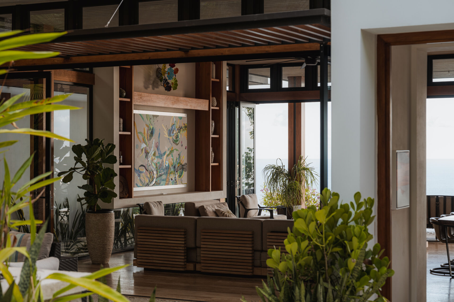 Warm wood, black-framed openings, lush greenery and colorful artwork define the relaxed living room at Tierra y Mar House, with ocean views beyond.