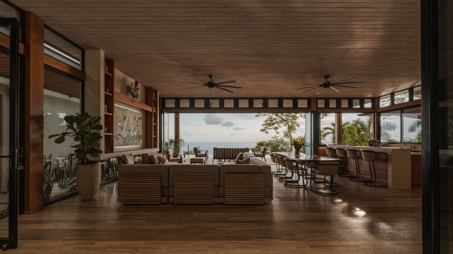 Warm wood, black-framed openings, lush greenery and colorful artwork define the relaxed living room at Tierra y Mar House, with ocean views beyond.