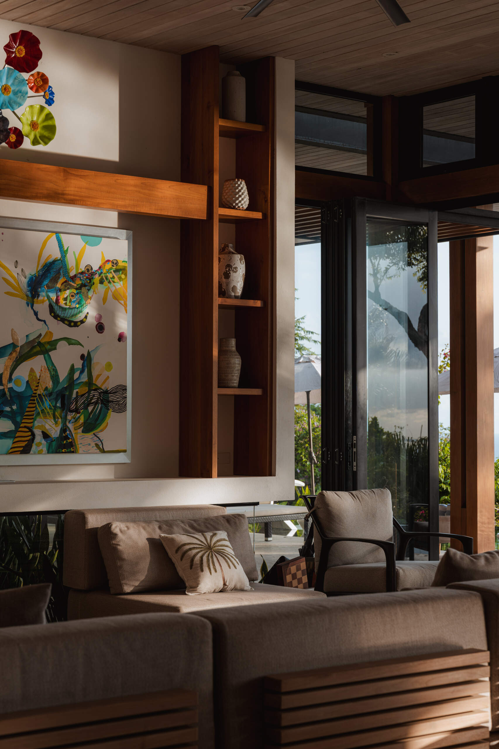 Warm wood, black-framed openings, lush greenery and colorful artwork define the relaxed living room at Tierra y Mar House, with ocean views beyond.