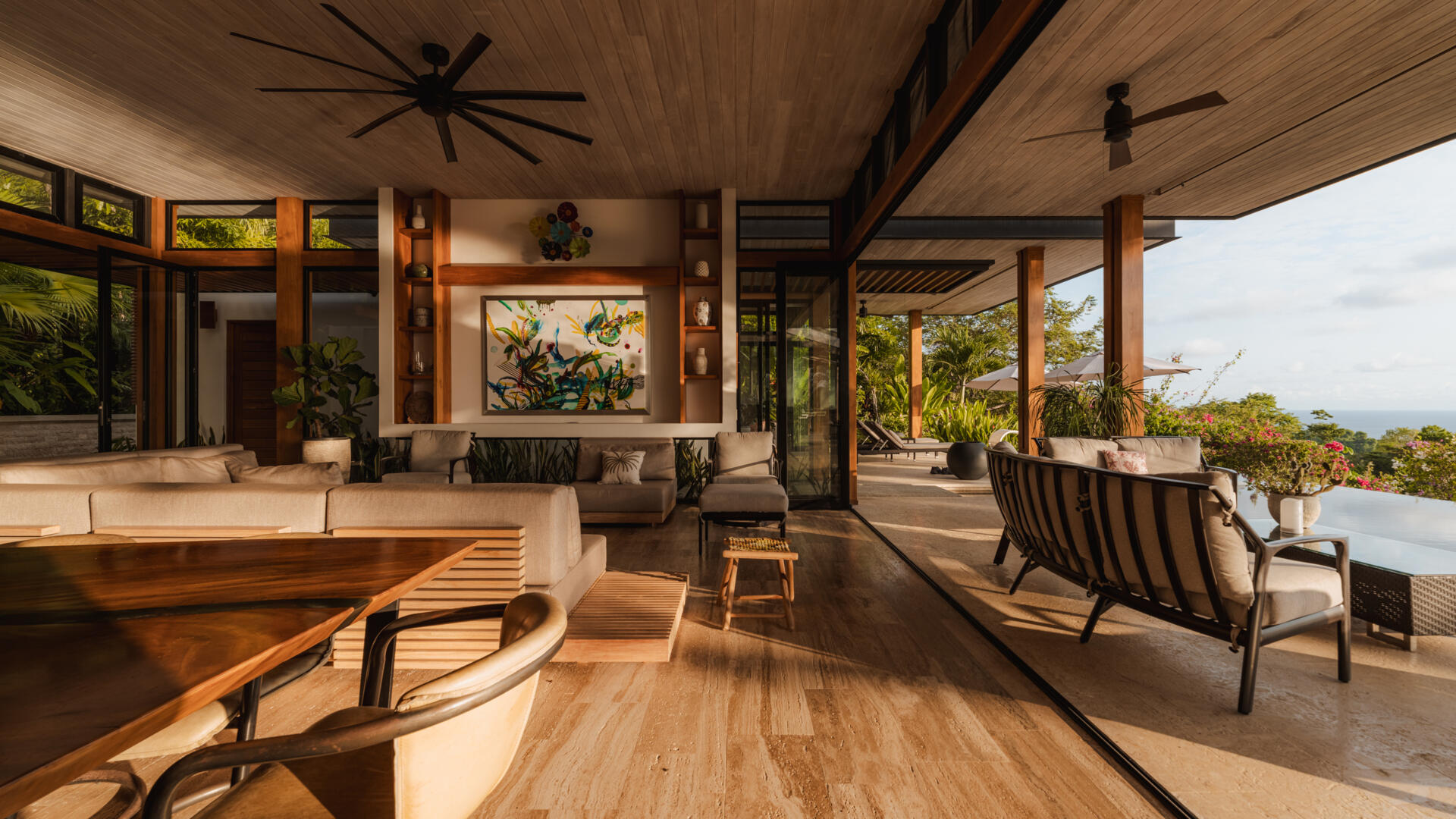 Warm wood, black-framed openings, lush greenery and colorful artwork define the relaxed living room at Tierra y Mar House, with ocean views beyond.