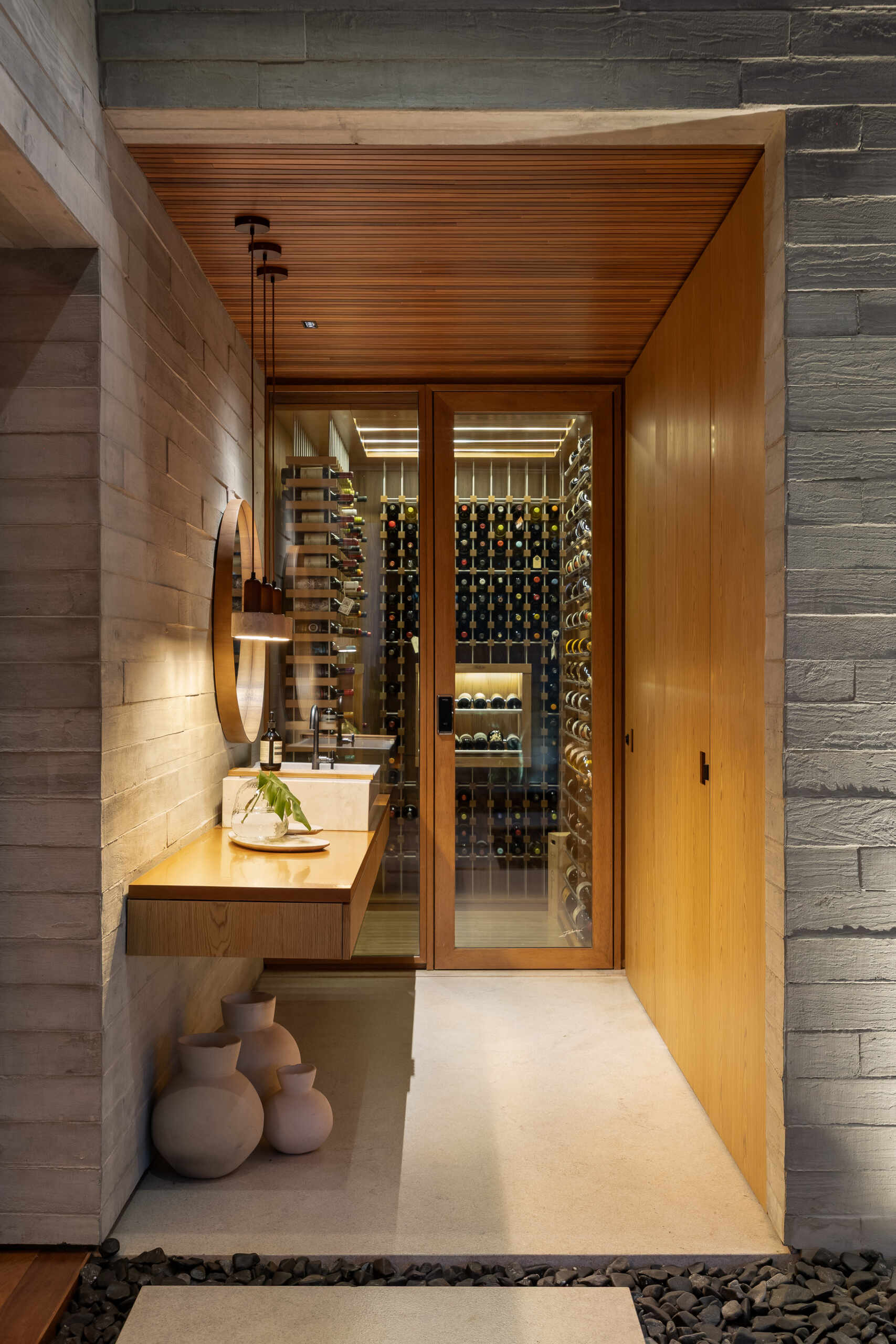 Near the wine cellar, warm wood and textured gray masonry frame a compact vanity area, while a glass-fronted wine cellar adds depth and visual interest to the passage.