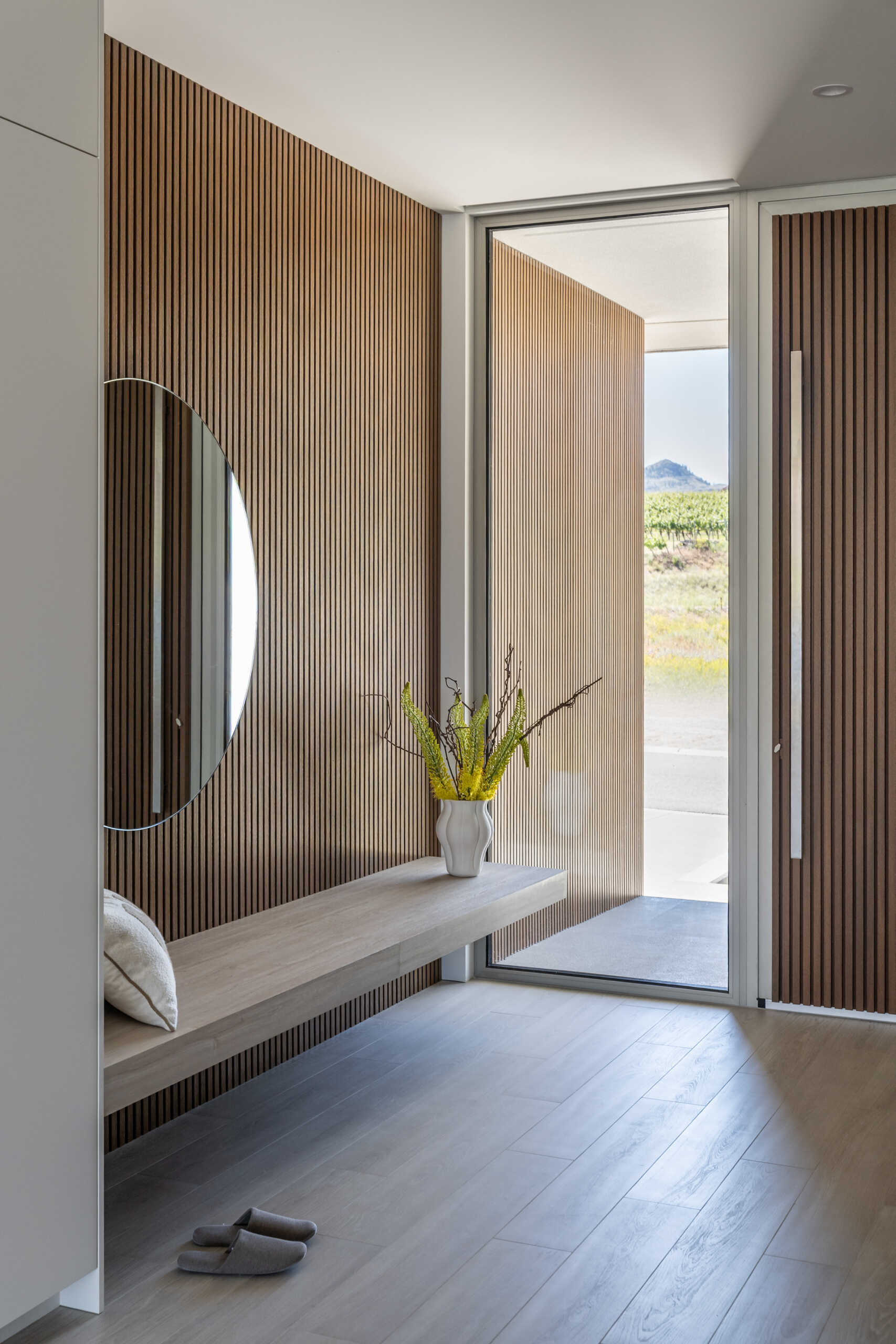 Step inside this modern lake house entryway, where warm vertical wood slats, a floating bench, round mirror, and tall glass create a bright and welcoming arrival with views toward the landscape.