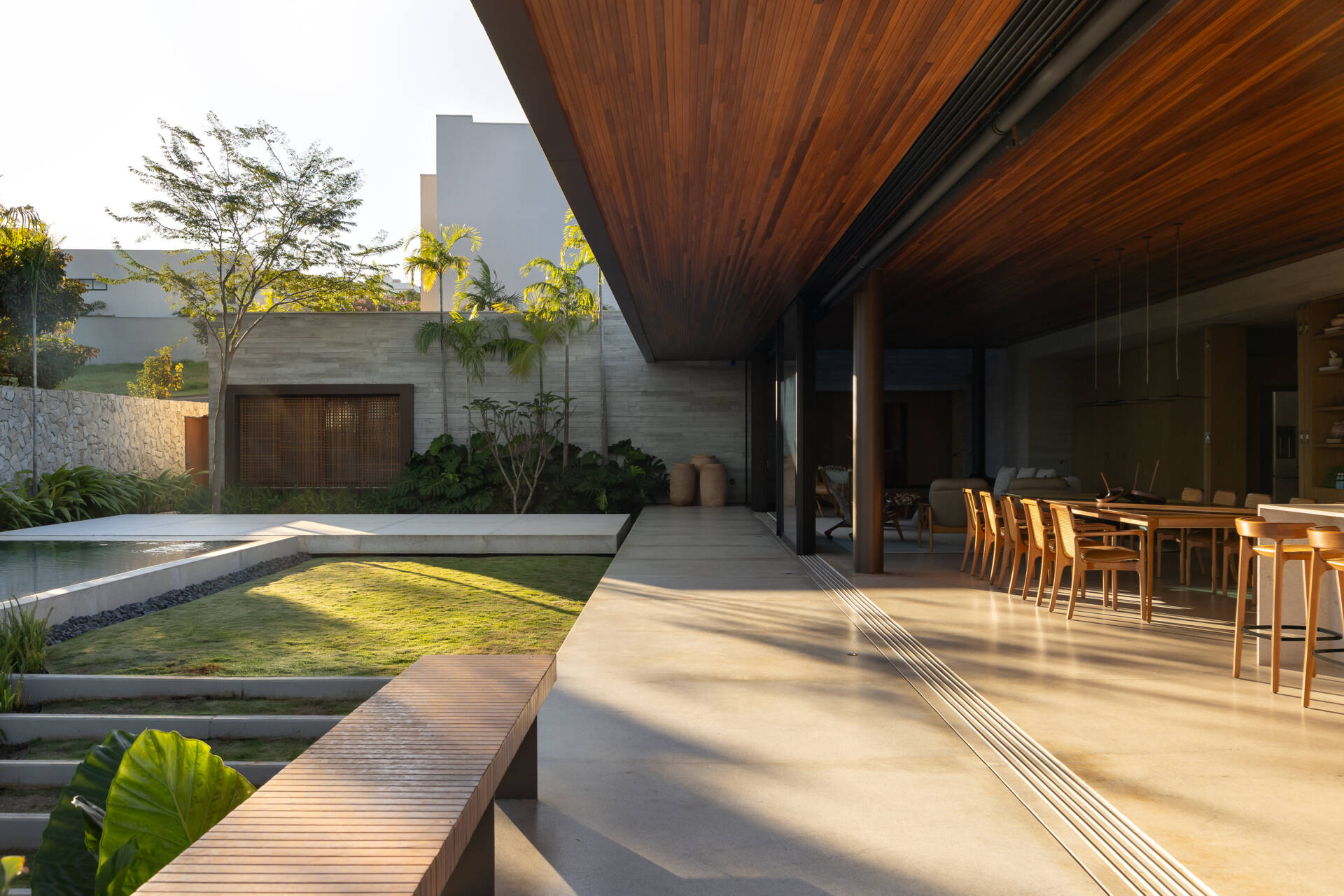 A deep wood-lined roof extends over Casa da Mata's open living and dining area, creating a sheltered transition to the garden. Concrete floors, dark columns, natural wood furniture, tropical planting and a reflecting pool complete the space.
