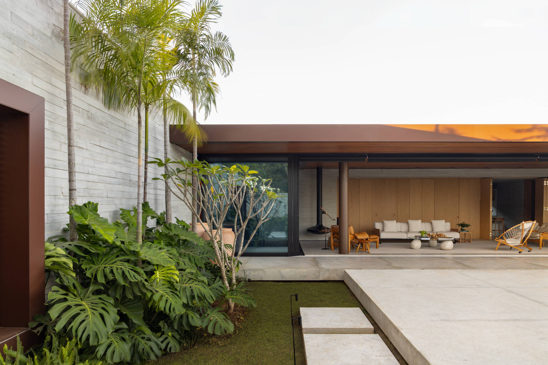 A deep wood-lined roof extends over Casa da Mata's open living and dining area, creating a sheltered transition to the garden. Concrete floors, dark columns, natural wood furniture, tropical planting and a reflecting pool complete the space.