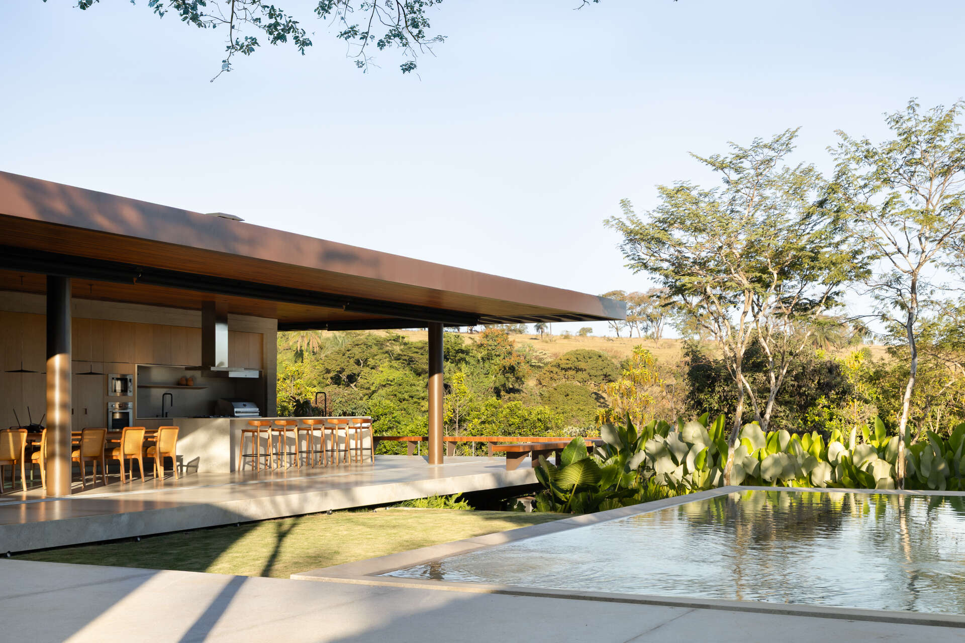 A deep wood-lined roof extends over Casa da Mata's open living and dining area, creating a sheltered transition to the garden. Concrete floors, dark columns, natural wood furniture, tropical planting and a reflecting pool complete the space.
